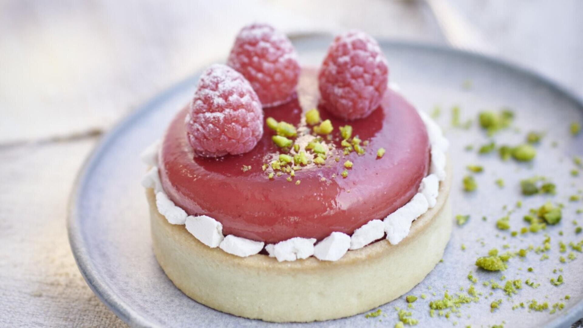 A round pastry topped with glossy raspberry glaze, three fresh raspberries, crushed pistachios, and white meringue pieces, served on a light blue plate with scattered pistachio crumbs.
