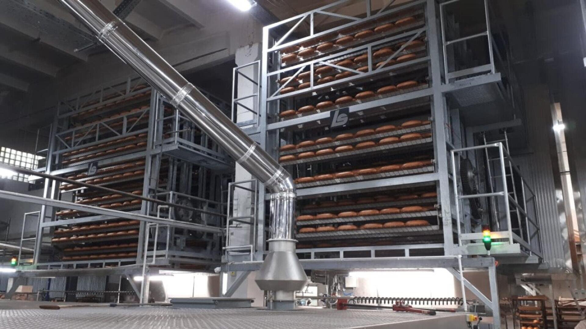 In einer geräumigen Fabrik stapeln sich große industrielle Bäckereigestelle, die mit Reihen von runden Broten gefüllt sind. Metallstrukturen und Rohre sind überall in der modernen, organisierten Anlage zu sehen.