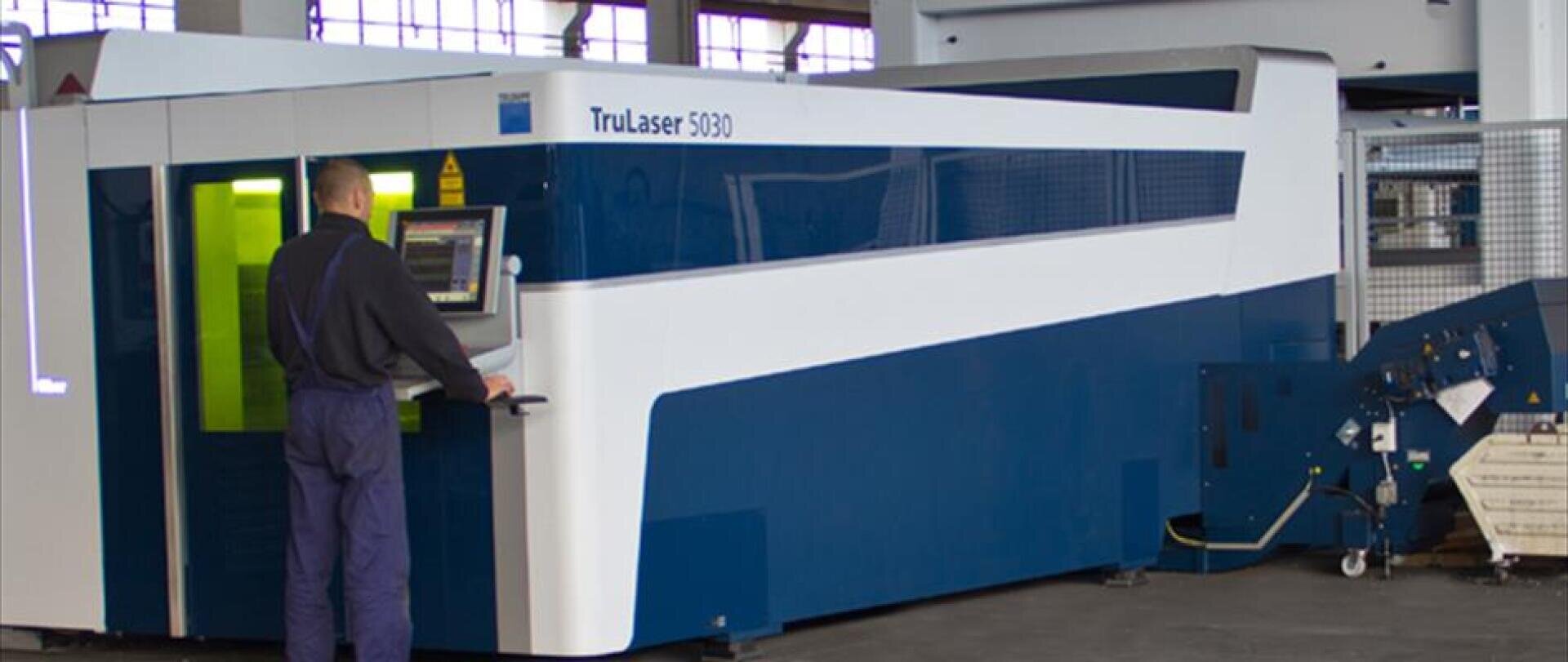 A worker operating a large blue and white TruLaser 5030 industrial laser cutting machine in a factory setting.