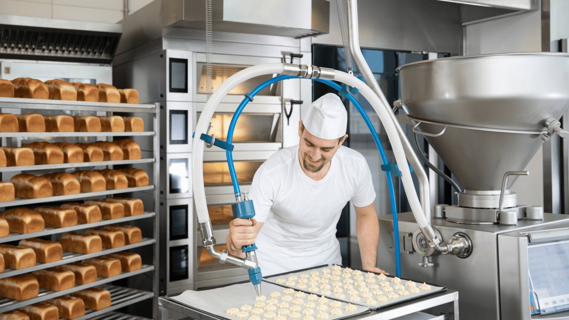 A baker in a white uniform and cap uses a machine to dispense dough onto a tray in a commercial bakery, with racks of bread loaves visible in the background.