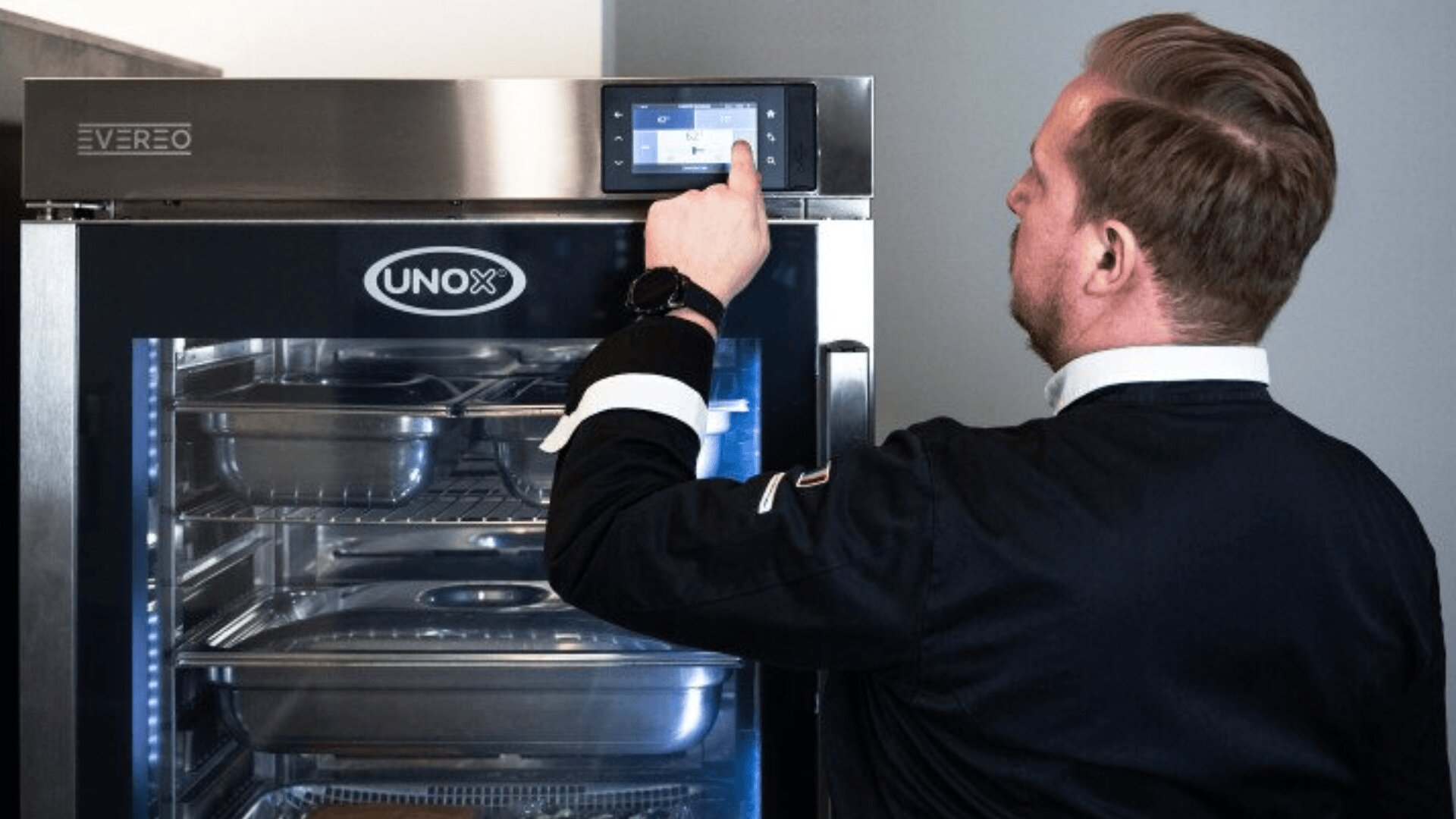 A person wearing a dark chefs jacket adjusts settings on a touchscreen control panel of a UNOX branded commercial kitchen appliance with metal trays inside.