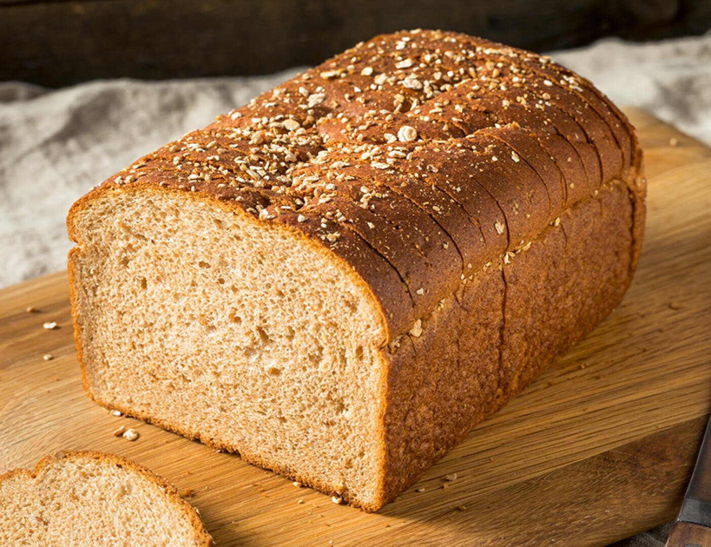 Auf einem hölzernen Schneidebrett liegt ein in Scheiben geschnittenes Vollkornbrot mit Haferflocken, von dem eine Scheibe teilweise abgeschnitten daneben liegt.