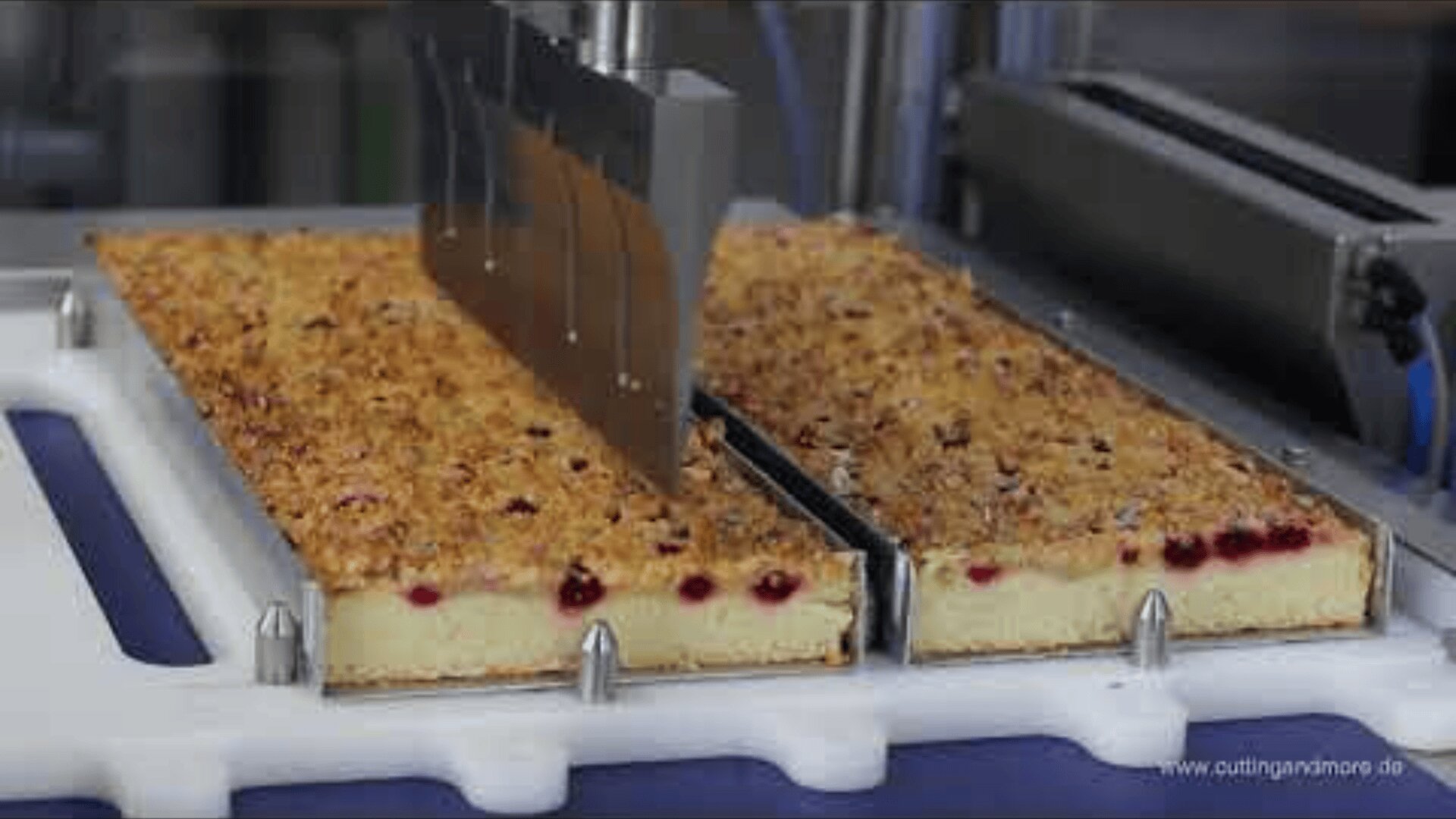 A machine with a large metal blade cuts two rectangular cakes topped with crumbs and red berries, placed on a white tray.