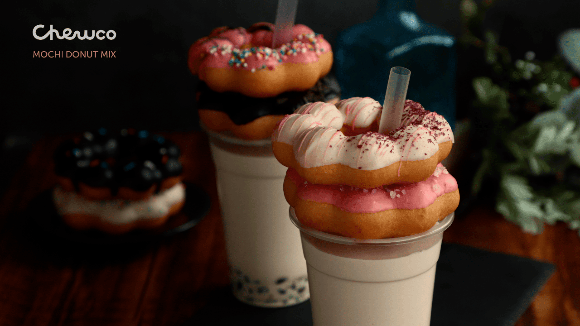 Zwei Bubble Tea-Getränke werden mit gestapelten, bunten Mochi-Donuts präsentiert. Ein Donut ist rosa mit Streuseln, der andere ist weiß mit rosa Zuckerguss. Der Hintergrund ist dunkel mit verschwommenem Grün und einer blauen Flasche.