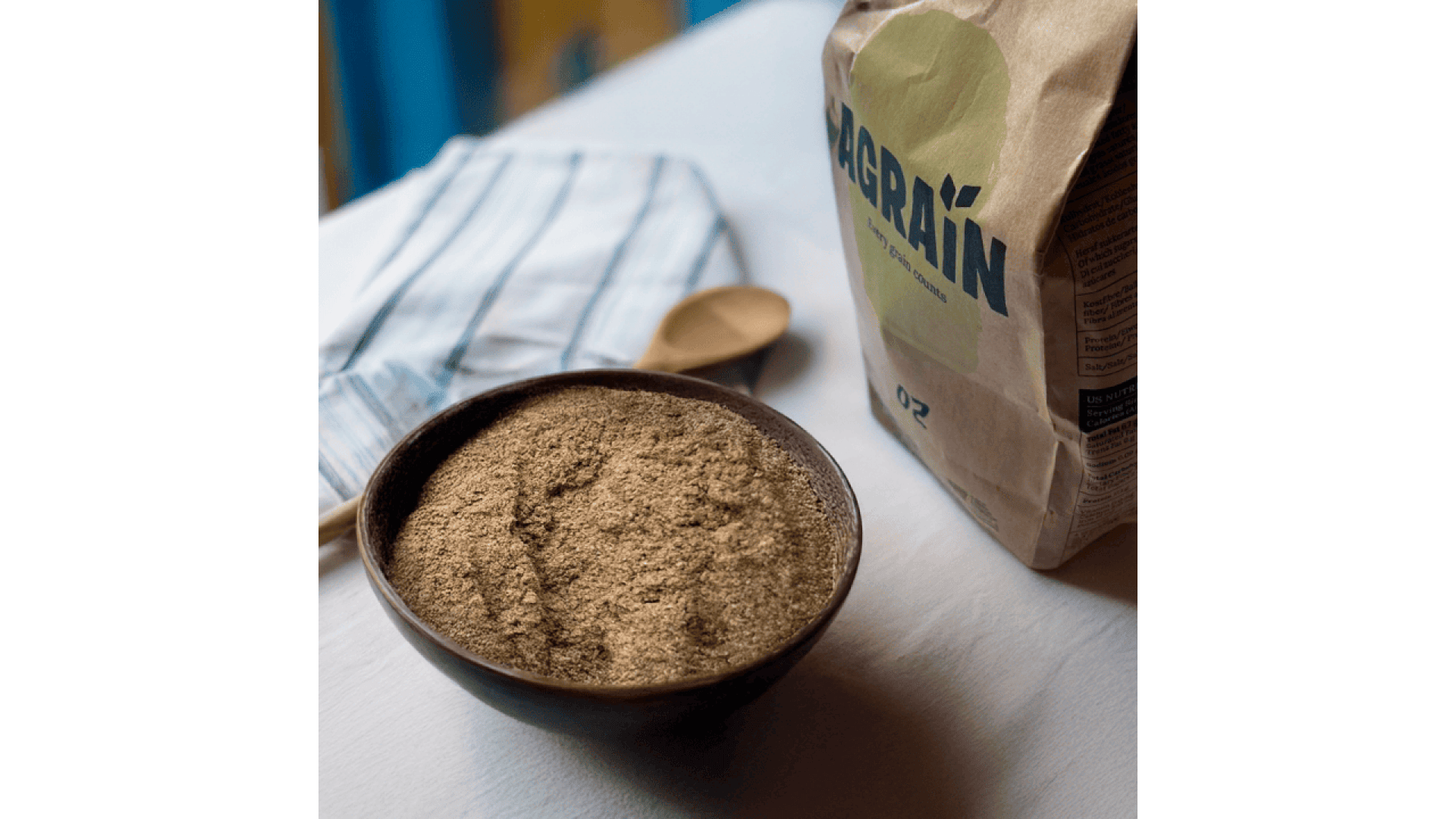 Auf einem Tisch steht eine mit braunem Mehl gefüllte Schüssel neben einer Papiertüte mit der Aufschrift „GAGRAIN“ und einem Holzlöffel auf einem gestreiften Tuch.