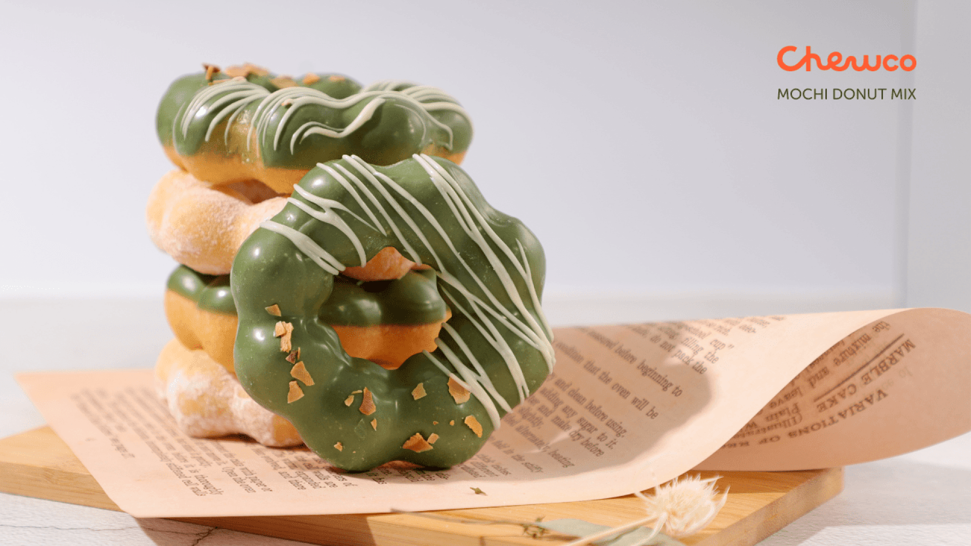 Drei Mochi Donuts mit grüner Glasur und weißem Nieselregen sind auf einem Holzbrett neben einem Blatt bedruckten Papiers gestapelt. Der Text Chewco MOCHI DONUT MIX steht in der oberen rechten Ecke.