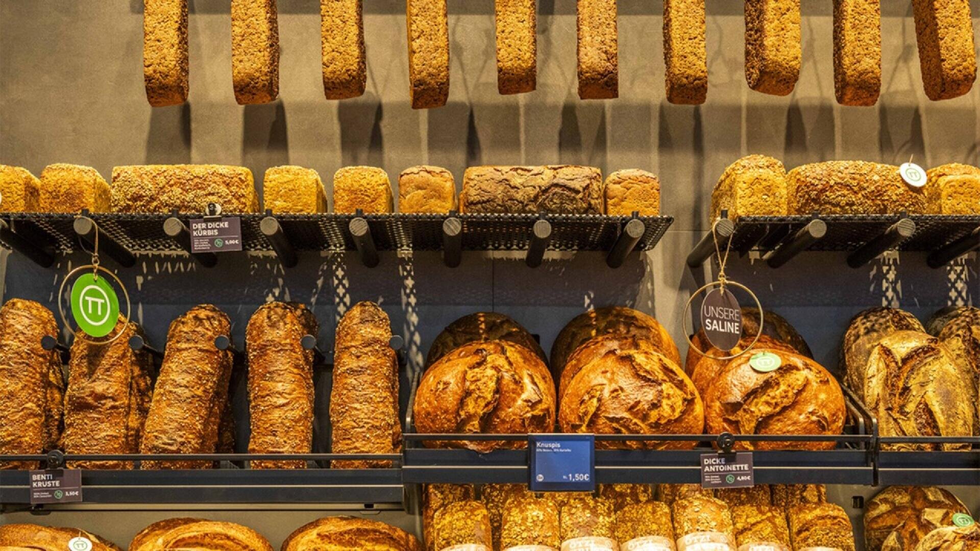 Loaves of various types of bread are displayed on shelves in a bakery, with several rows of crusty, rustic bread arranged neatly, some hanging and others stacked.