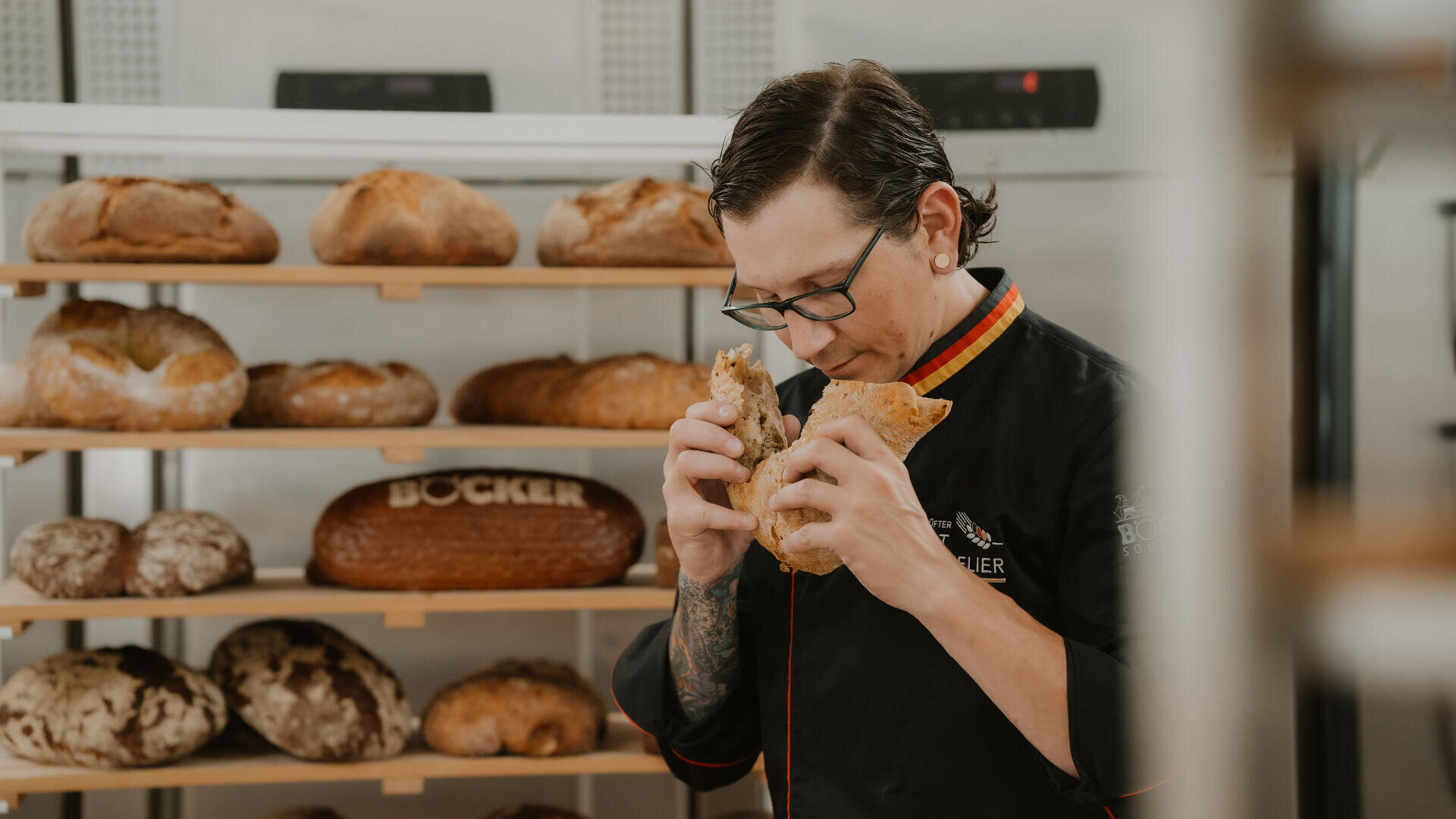 Ein Bäcker mit Brille und schwarzem Kittel riecht an einem Stück Brot in einer Bäckerei, im Hintergrund stehen Regale mit verschiedenen Brotsorten.