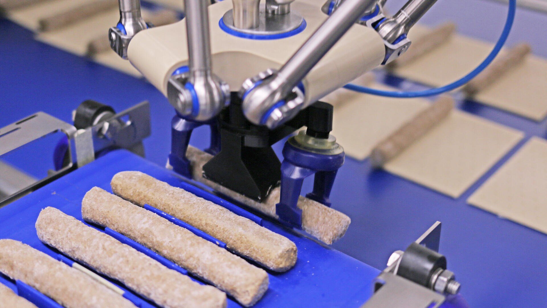 A robotic arm with a gripper picks up cylindrical food items from a blue conveyor belt in a manufacturing facility, with rectangular crackers in the background.