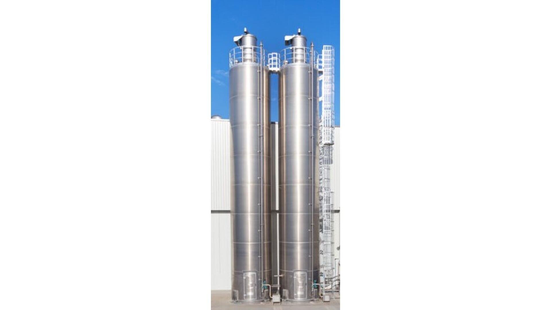 Zwei hohe, zylindrische Metallsilos mit Leitern und Geländern stehen nebeneinander vor einem weißen Industriegebäude unter blauem Himmel.