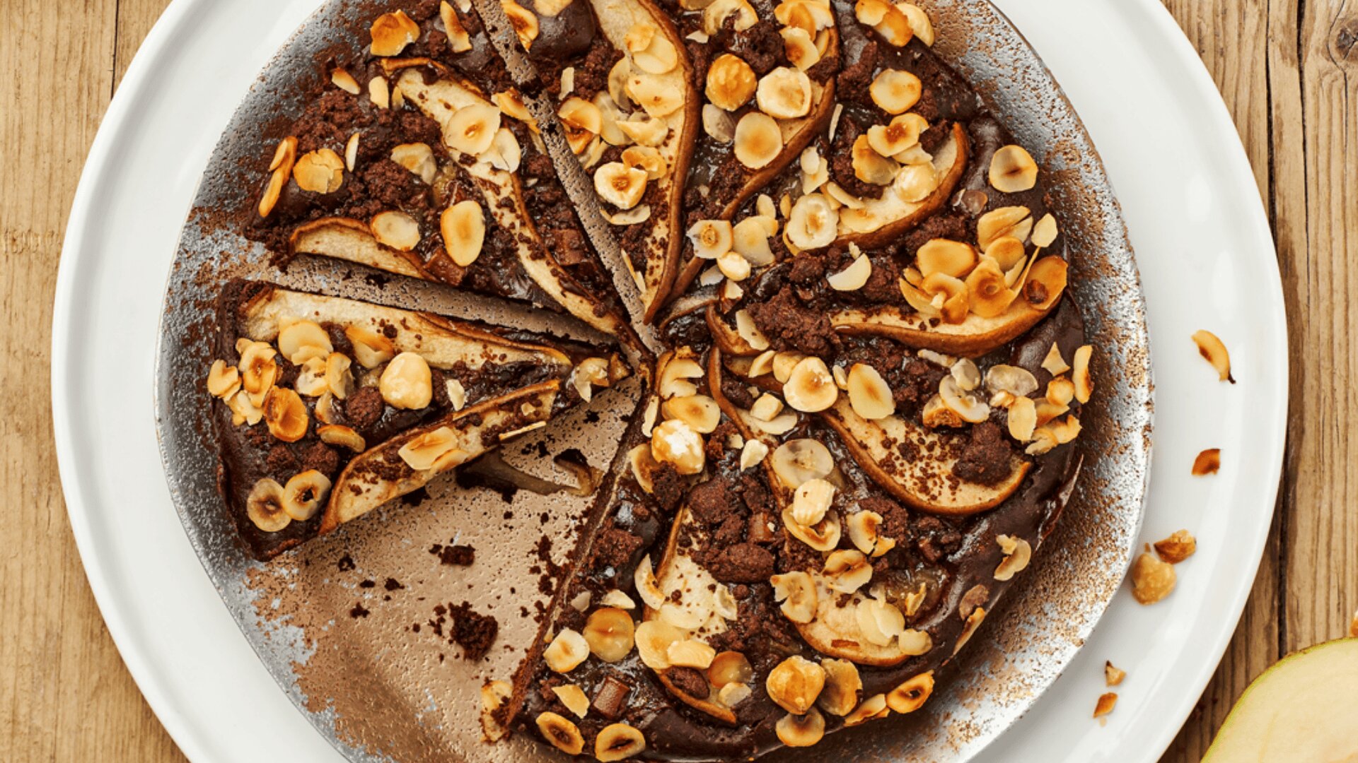 A top view of a sliced chocolate cake topped with pear slices and chopped hazelnuts, served on a white plate on a wooden surface.