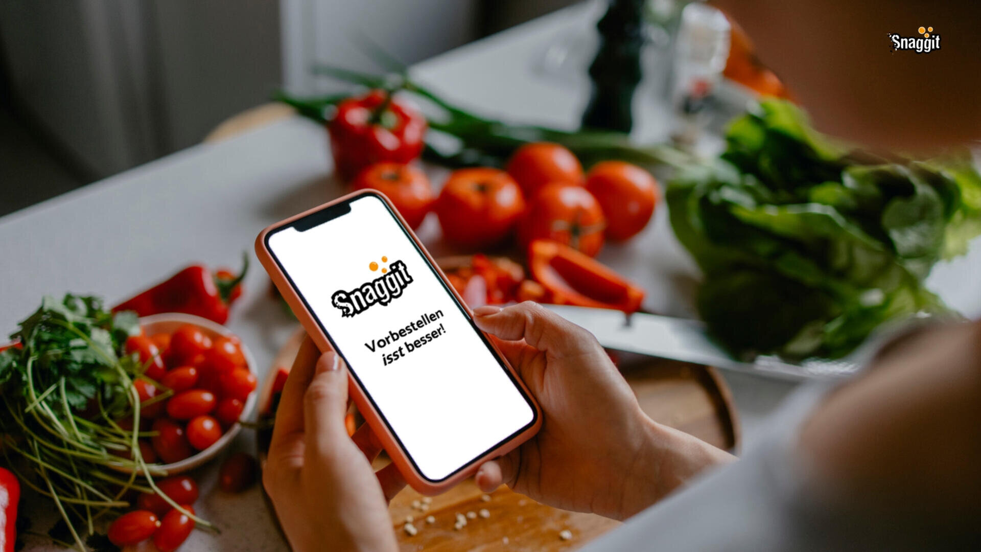 Eine Person hält ein Smartphone mit der Snaggit-App und dem deutschen Text „Vorbestellen ist besser!“ in einer Küche mit frischem Gemüse wie Tomaten und Salat auf einem Schneidebrett.