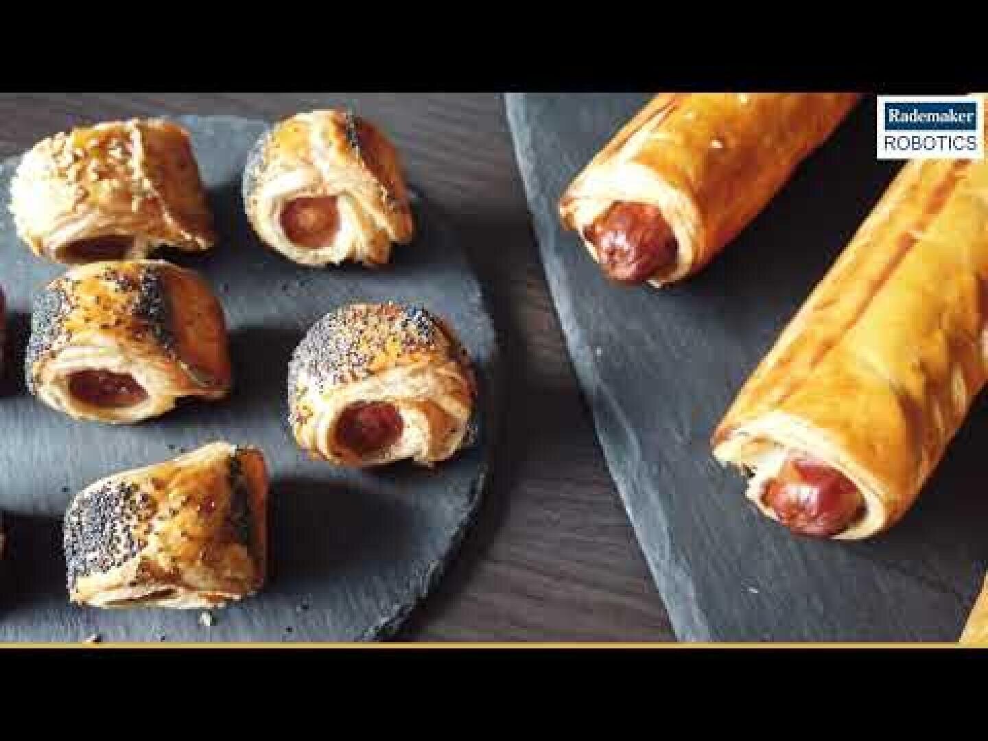 Verschiedene gebackene Wurstsemmeln, einige mit Mohn, werden auf dunklen Servierplatten präsentiert. Das Gebäck ist goldbraun und flockig, und es sind ganze und geschnittene Brötchen zu sehen.