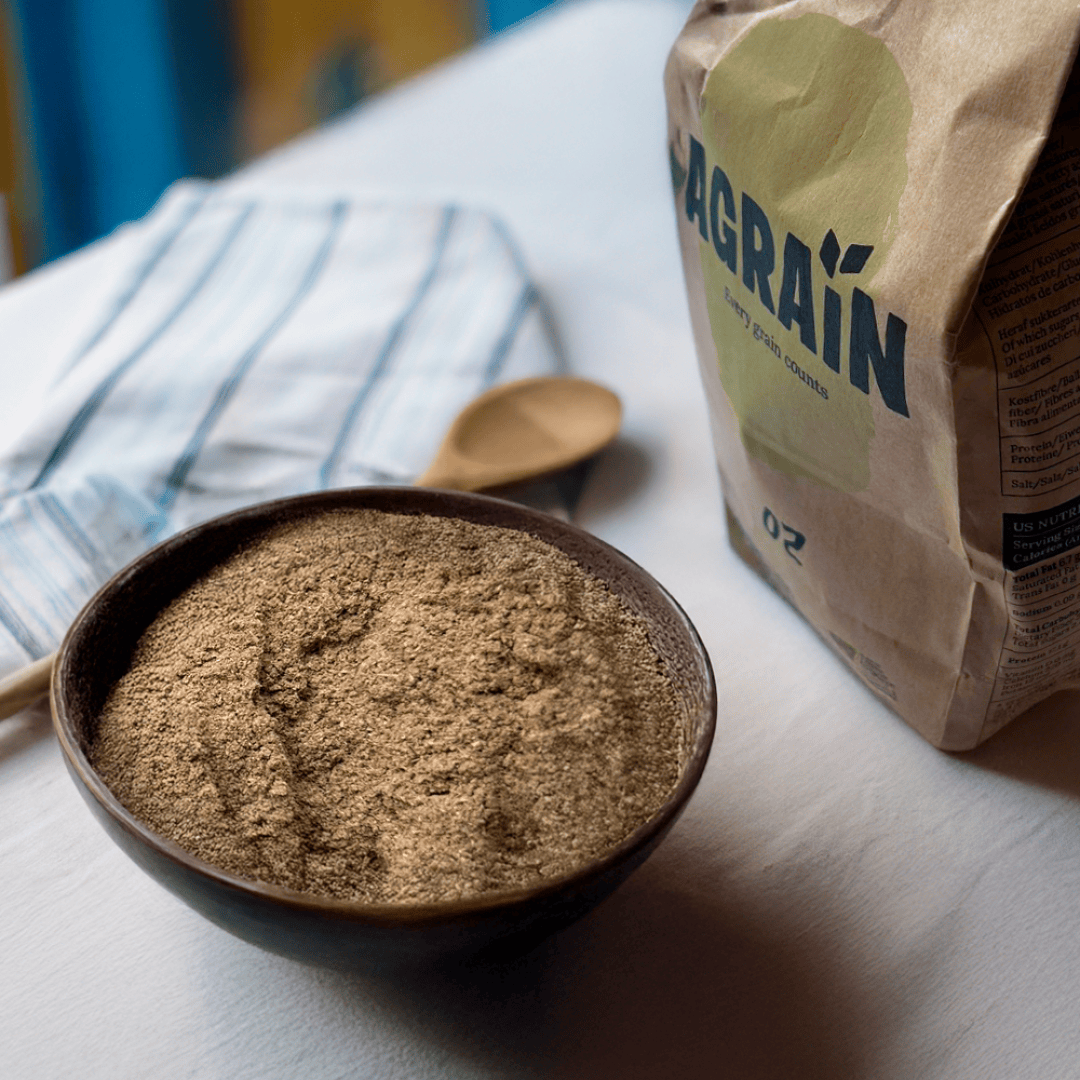 A brown bowl filled with flour sits on a white table next to a paper bag labeled GRAIN. A striped cloth and a wooden spoon are in the background.