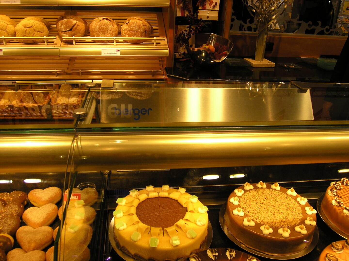 Eine Bäckereivitrine, gefüllt mit verschiedenen Kuchen, Gebäck und Backwaren, darunter runde Kuchen mit Zuckerguss und herzförmige Kekse, alles hell beleuchtet und ordentlich auf Regalen angeordnet.