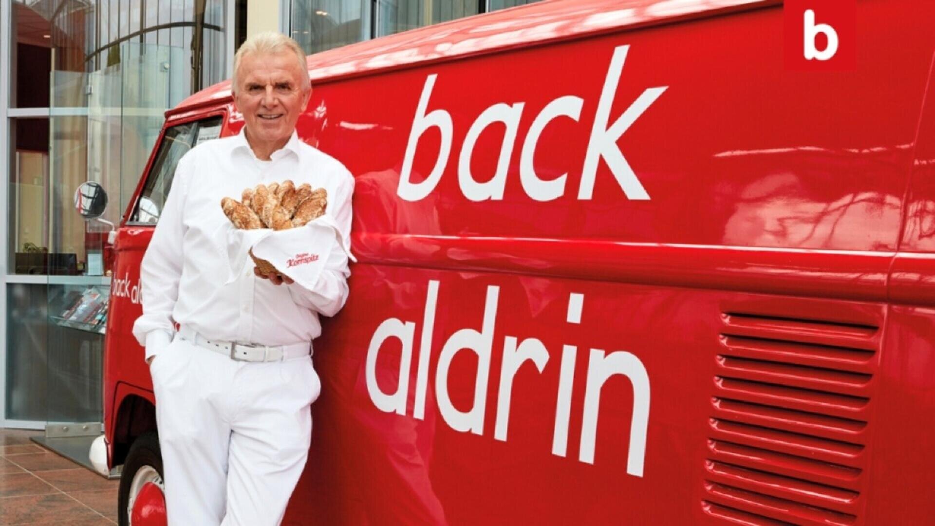 An older man in a white outfit holds a basket of baguettes and leans against a red van with the words back aldrin written on the side. The scene is in front of a glass building.
