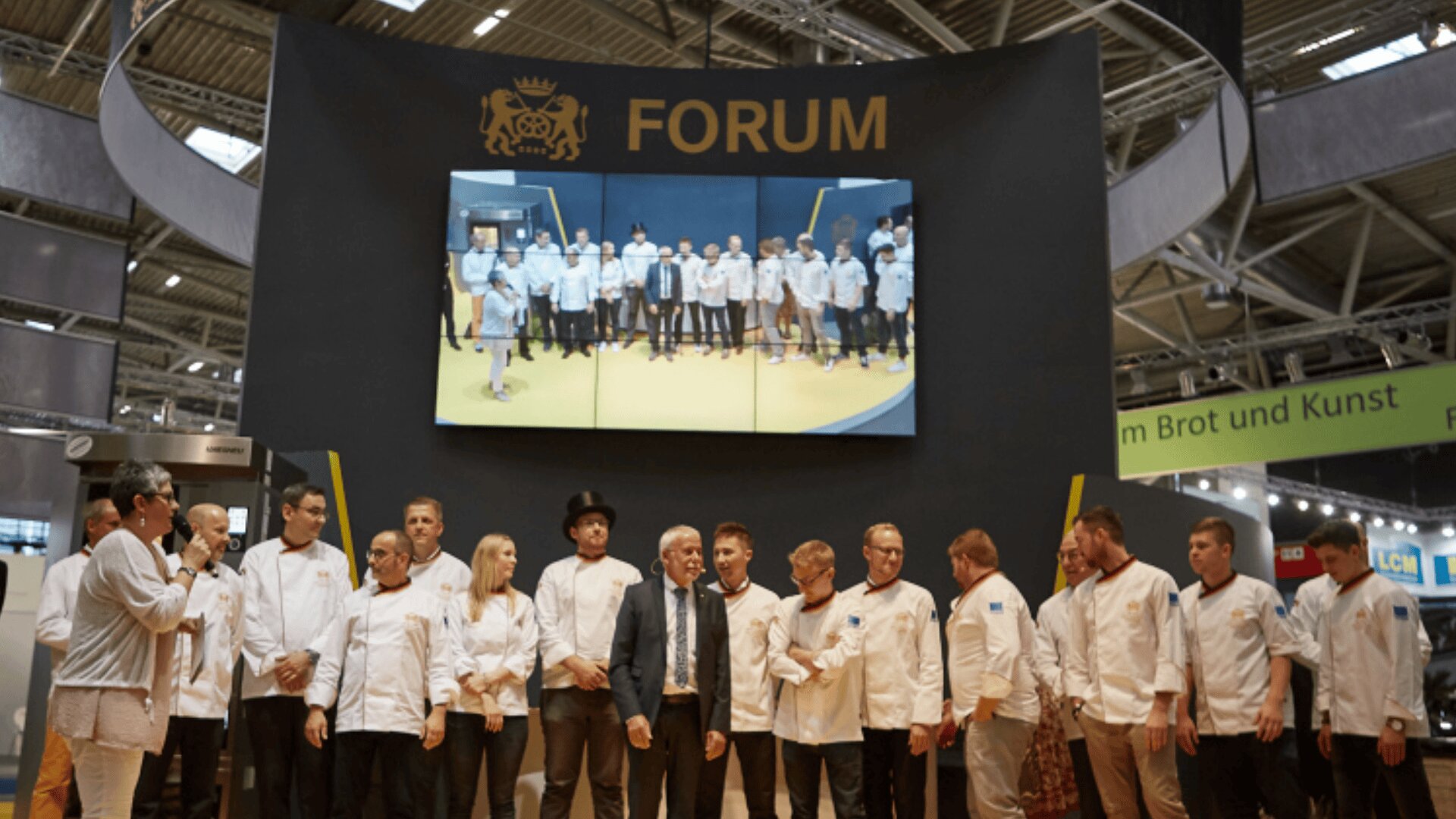 Eine Gruppe von Köchen und Beamten steht auf der Bühne unter einem großen FORUM-Schild, über dem ein Bildschirm mit einem Foto von weiteren Köchen zu sehen ist. Die Veranstaltung scheint in einem Kongresszentrum stattzufinden.