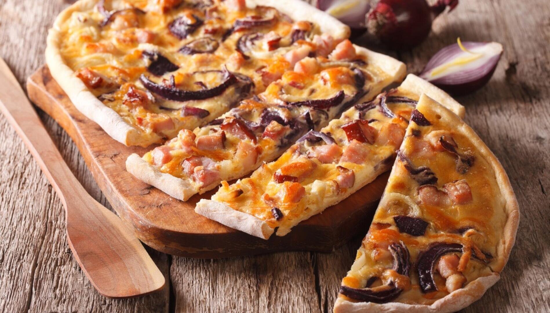Eine rechteckige Pizza mit dünnem Boden, belegt mit Speck, roten Zwiebeln und Käse, wird in Scheiben geschnitten und auf einem Holzbrett serviert, im Hintergrund ein Holzspatel und rohe Zwiebeln.