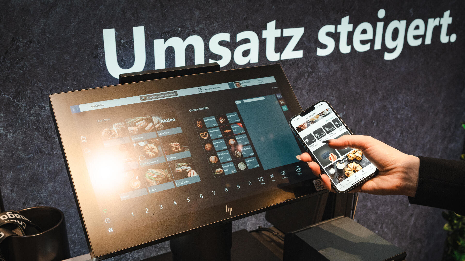 Eine Person hält ein Smartphone, auf dem eine Speisekarte angezeigt wird, vor einem Touchscreen-Kassensystem, das ähnliche Lebensmitteloptionen anzeigt. Im Hintergrund sind die deutschen Worte "Umsatz steigt" zu sehen.