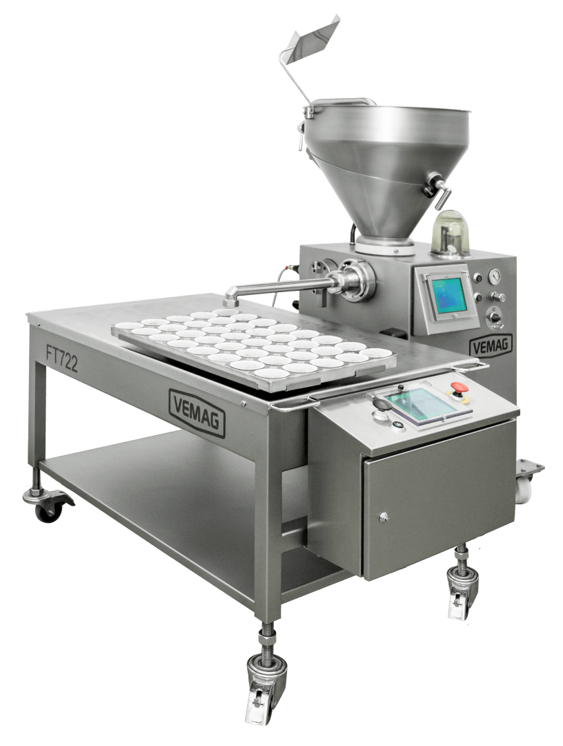 Eine industrielle Lebensmittelverarbeitungsmaschine aus Edelstahl mit einem Touchscreen-Bedienfeld, einem Trichter und einem Förderband, das Schalen mit Lebensmitteln aufnimmt, mit der Bezeichnung VEMAG FT722, auf Rollen für Mobilität.