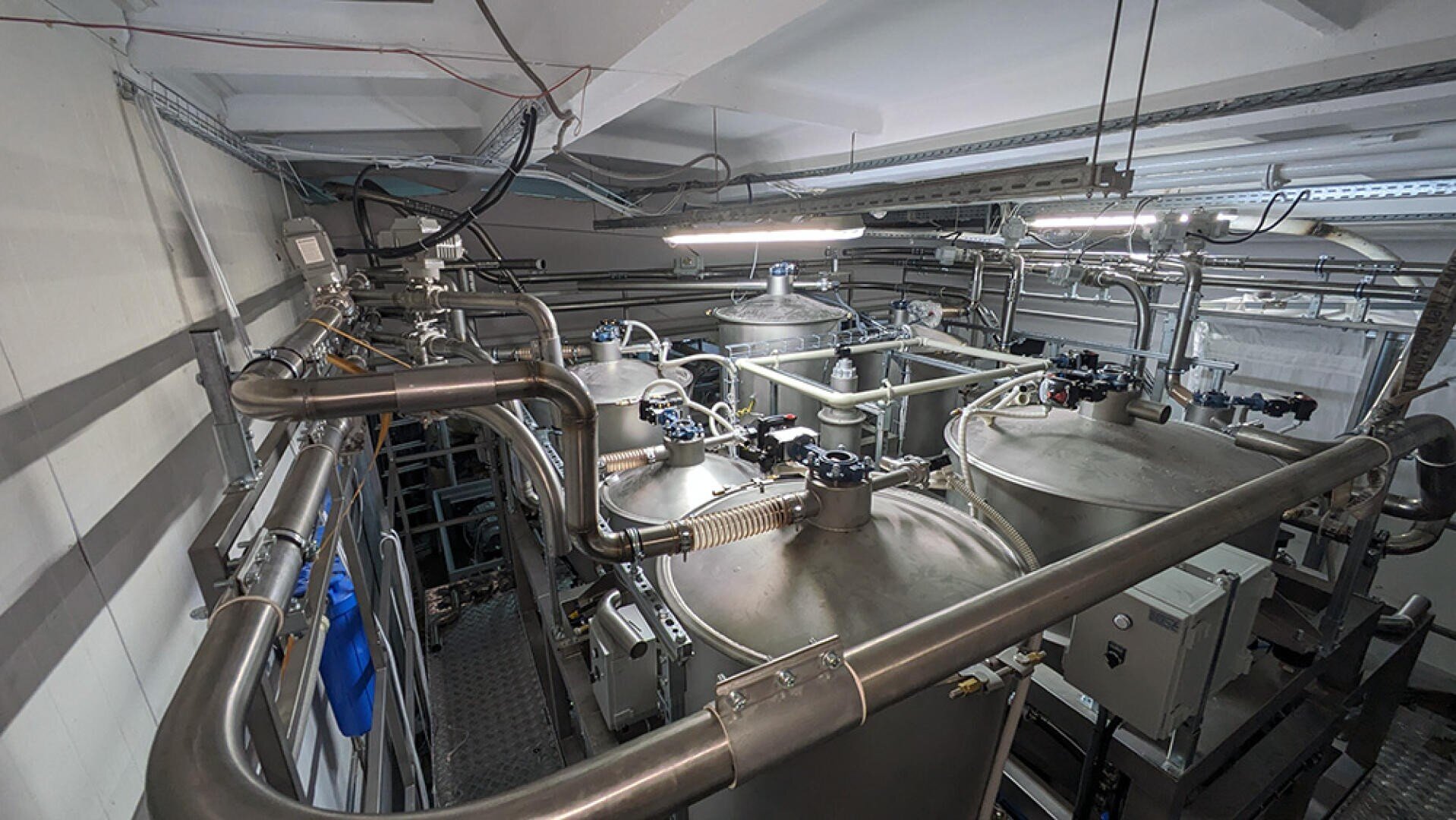 Large metal tanks and pipes in an industrial facility, possibly used for water treatment or chemical processing, with overhead lighting and various valves and control panels visible.