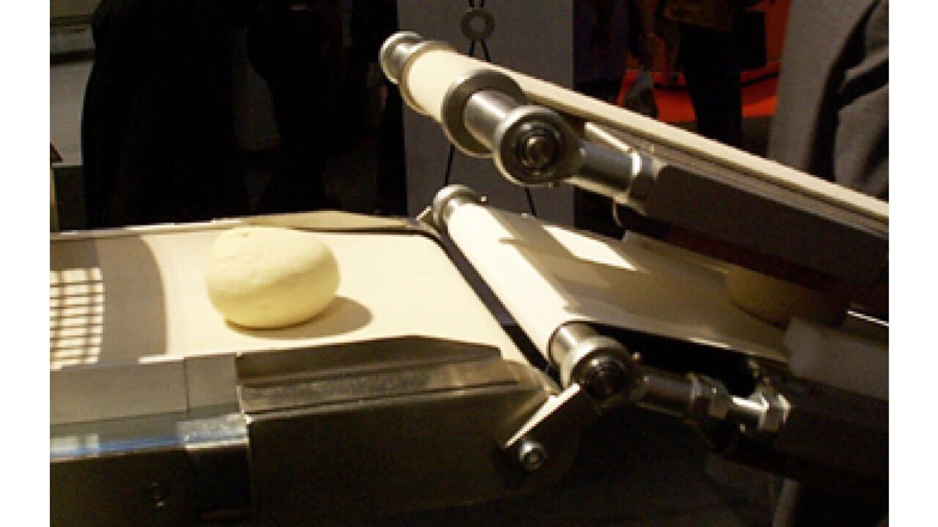 A close-up of an industrial conveyor belt system transporting a round dough ball through a metal and roller mechanism in a factory setting.
