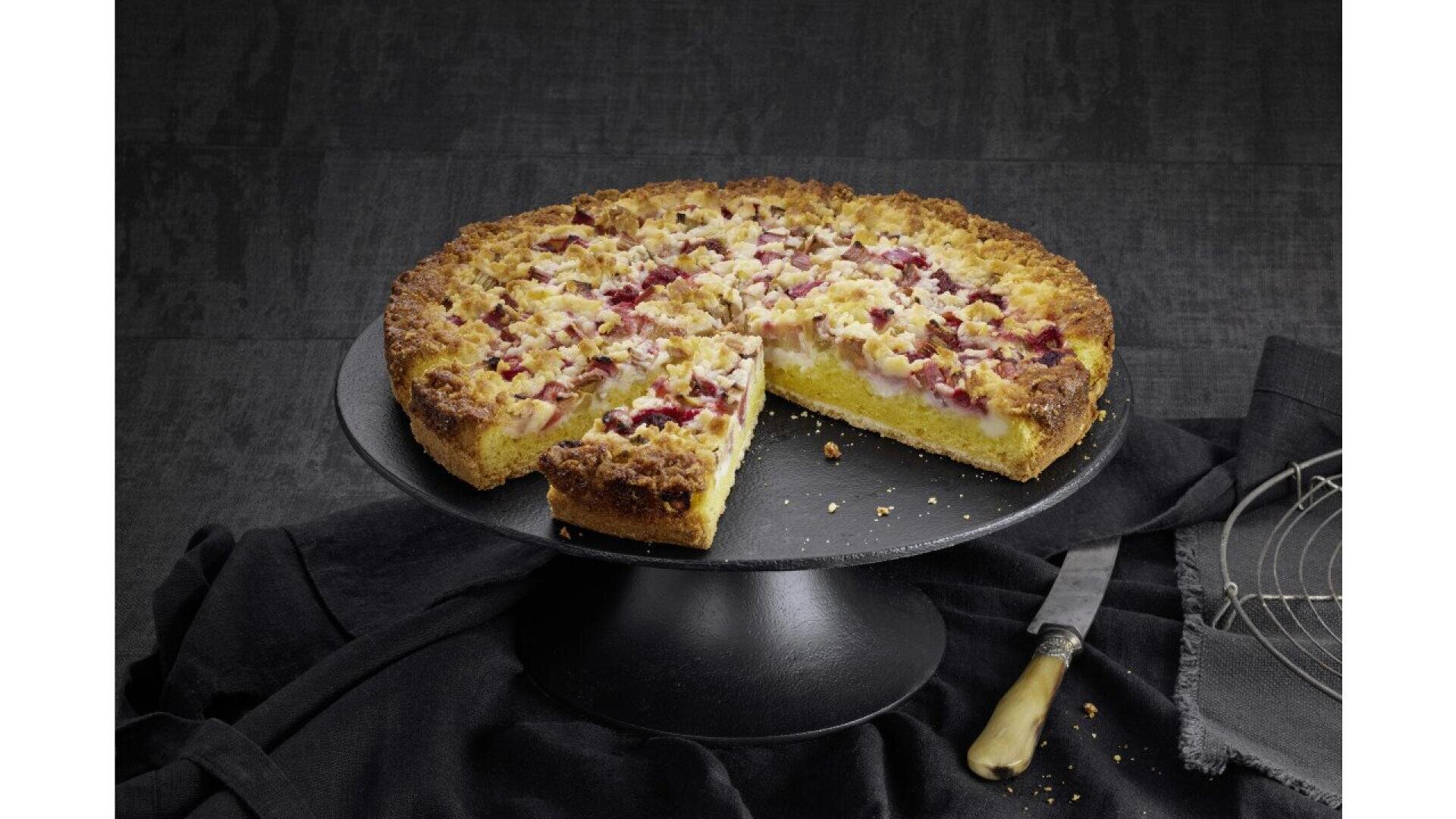 A round fruit crumble cake with a golden crust and red fruit pieces is displayed on a black cake stand. One slice has been cut and slightly removed, and a knife rests beside the stand on a dark surface.
