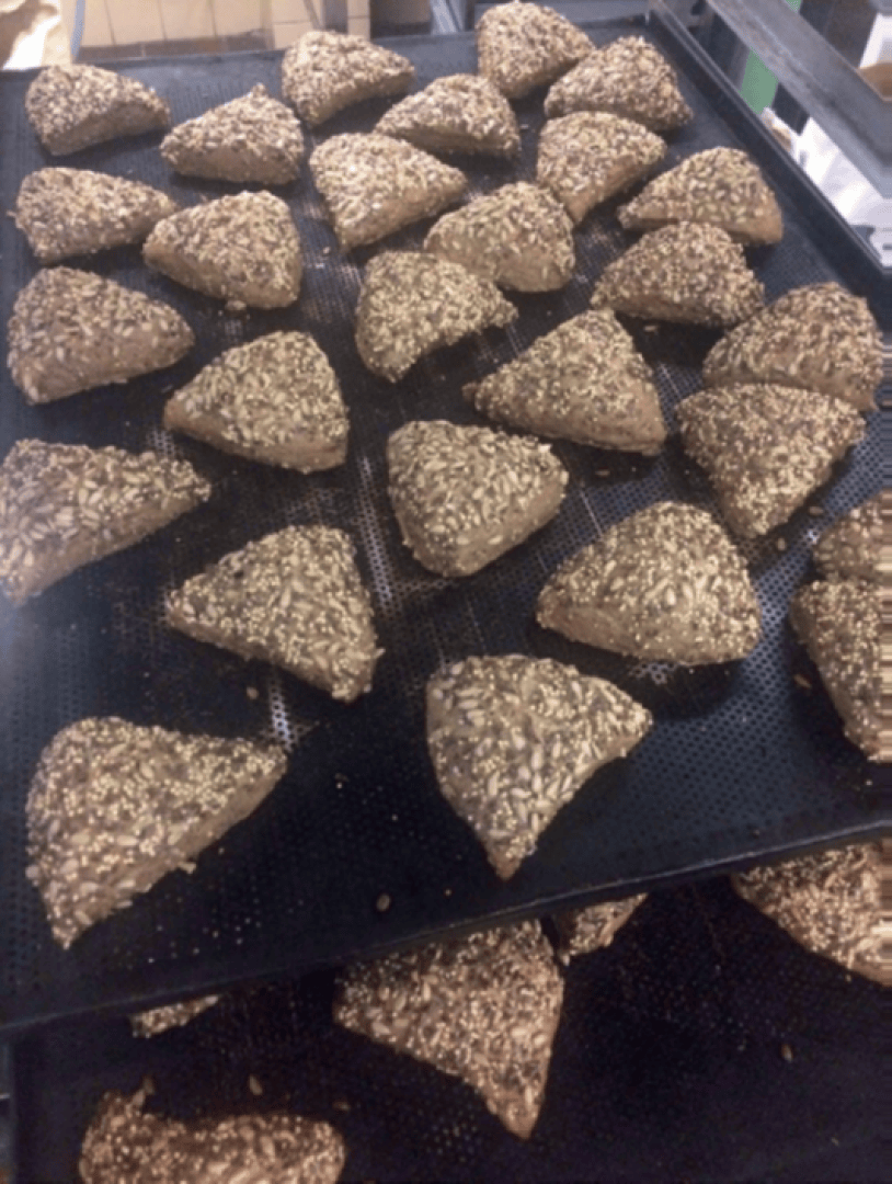 Mehrere dreieckige, braune Brötchen mit Körnern liegen in Reihen auf einem schwarzen Backblech, bereit zum Backen.