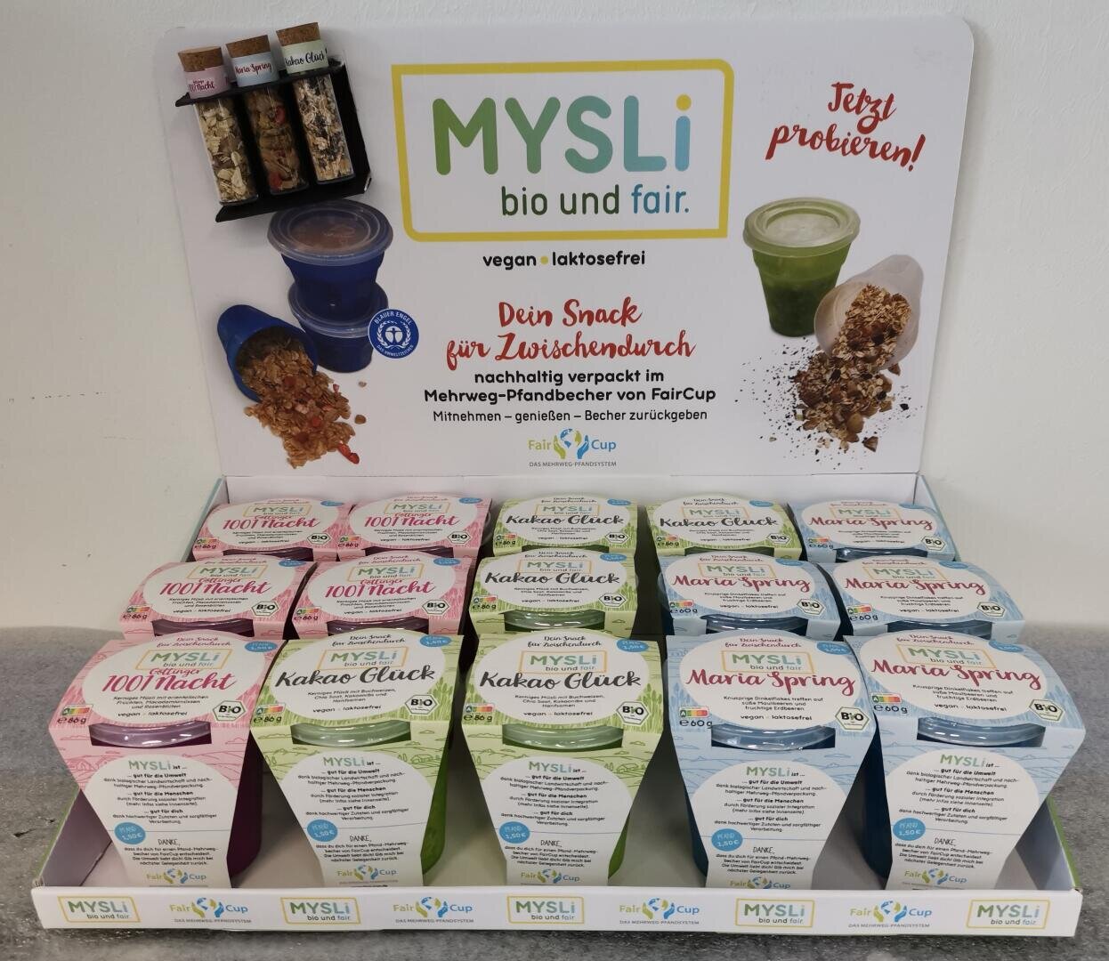 Eine Auslage von MYSLI Bio- und Fair-Müsli-Snacks in bunten Kartons, in denen vier Sorten in Reihen angeordnet sind. Dahinter befinden sich Produktproben in kleinen Gläsern, zwei Joghurtbecher und ein deutscher Werbetext.