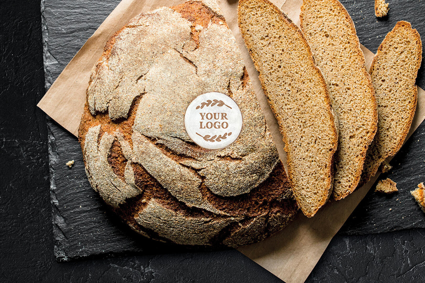 Ein runder Laib knusprigen Brotes mit mehreren geschnittenen Scheiben, die auf Pergamentpapier auf einer dunklen Schieferoberfläche präsentiert werden. Ein kreisförmiges essbares Etikett mit der Aufschrift YOUR LOGO befindet sich oben auf dem Brot.