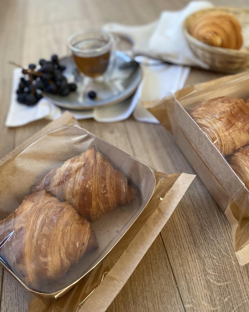 Two croissants in a clear plastic box sit on a wooden table. In the background, there is a cup of coffee, a bunch of grapes, and a basket with more croissants on a white napkin.