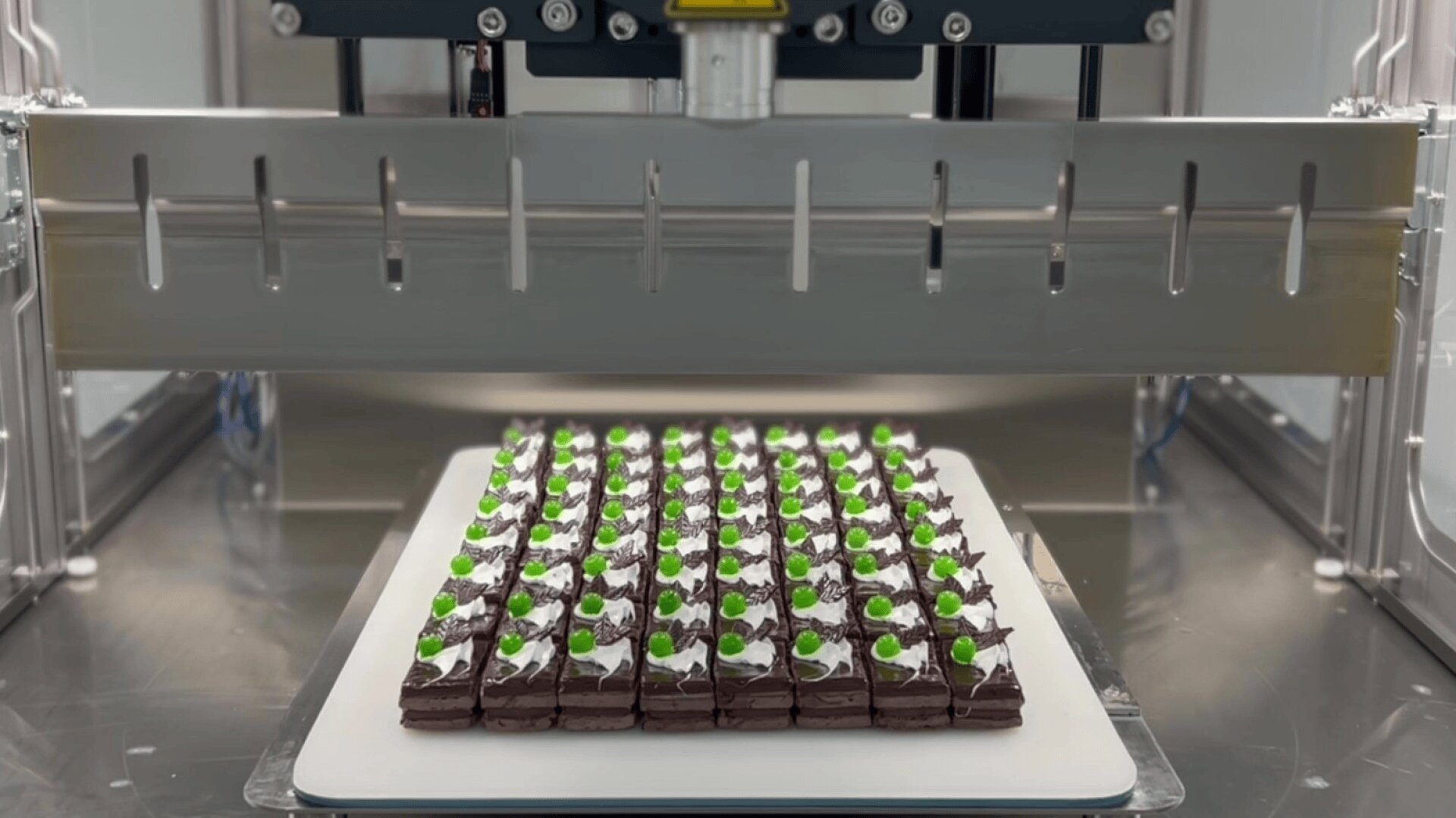A machine is positioned above a tray of neatly arranged chocolate desserts, each topped with white cream and a green garnish, ready for processing or decoration in a modern food production setting.