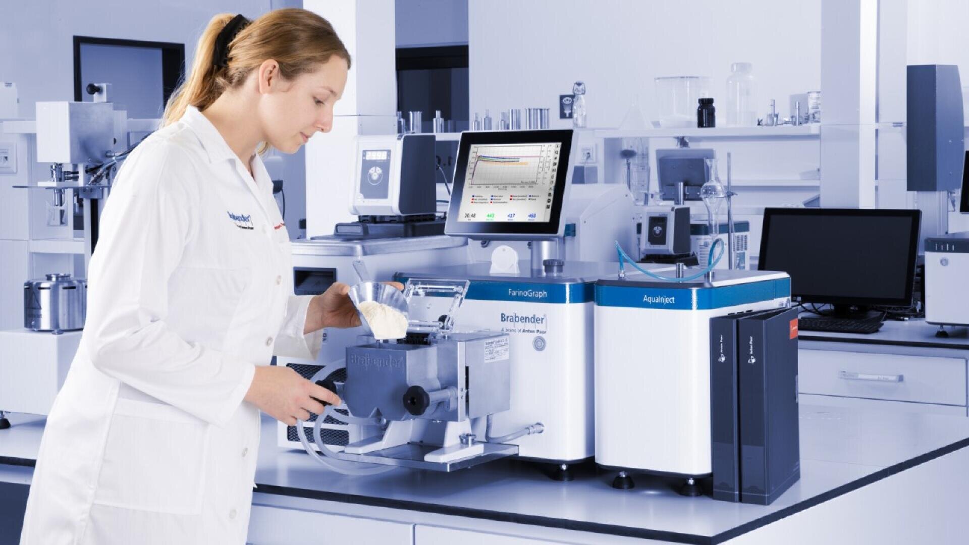 A woman in a white lab coat works in a laboratory, pouring powder into a machine with a digital display and various scientific equipment surrounding her on the counter.
