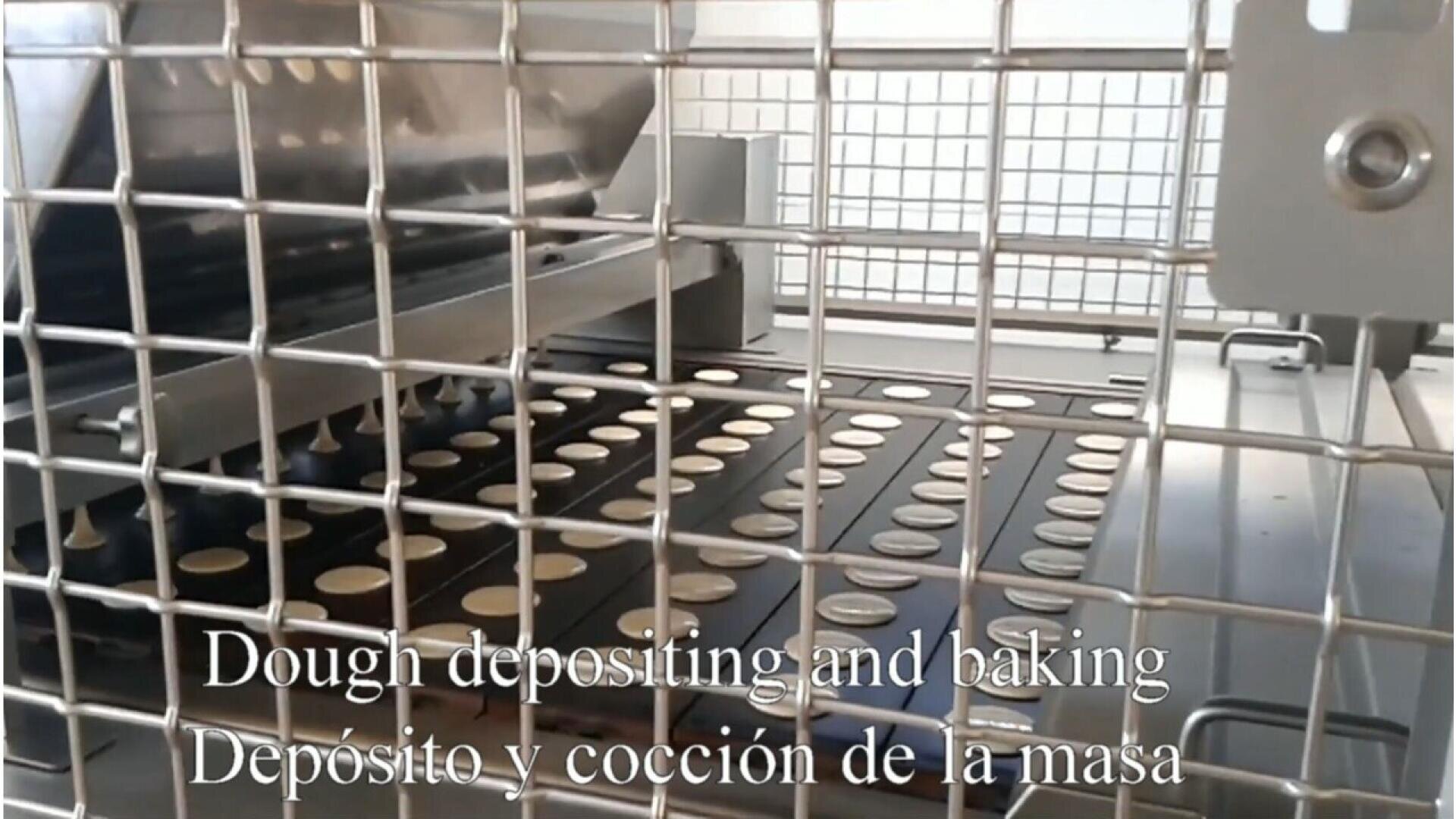Eine Teigablage- und Backmaschine formt kleine runde Teigstücke auf einem Tablett, gesehen durch ein Metallschutzgitter. Der Text am unteren Rand lautet "Dough depositing and baking / Depósito y cocción de la masa.".