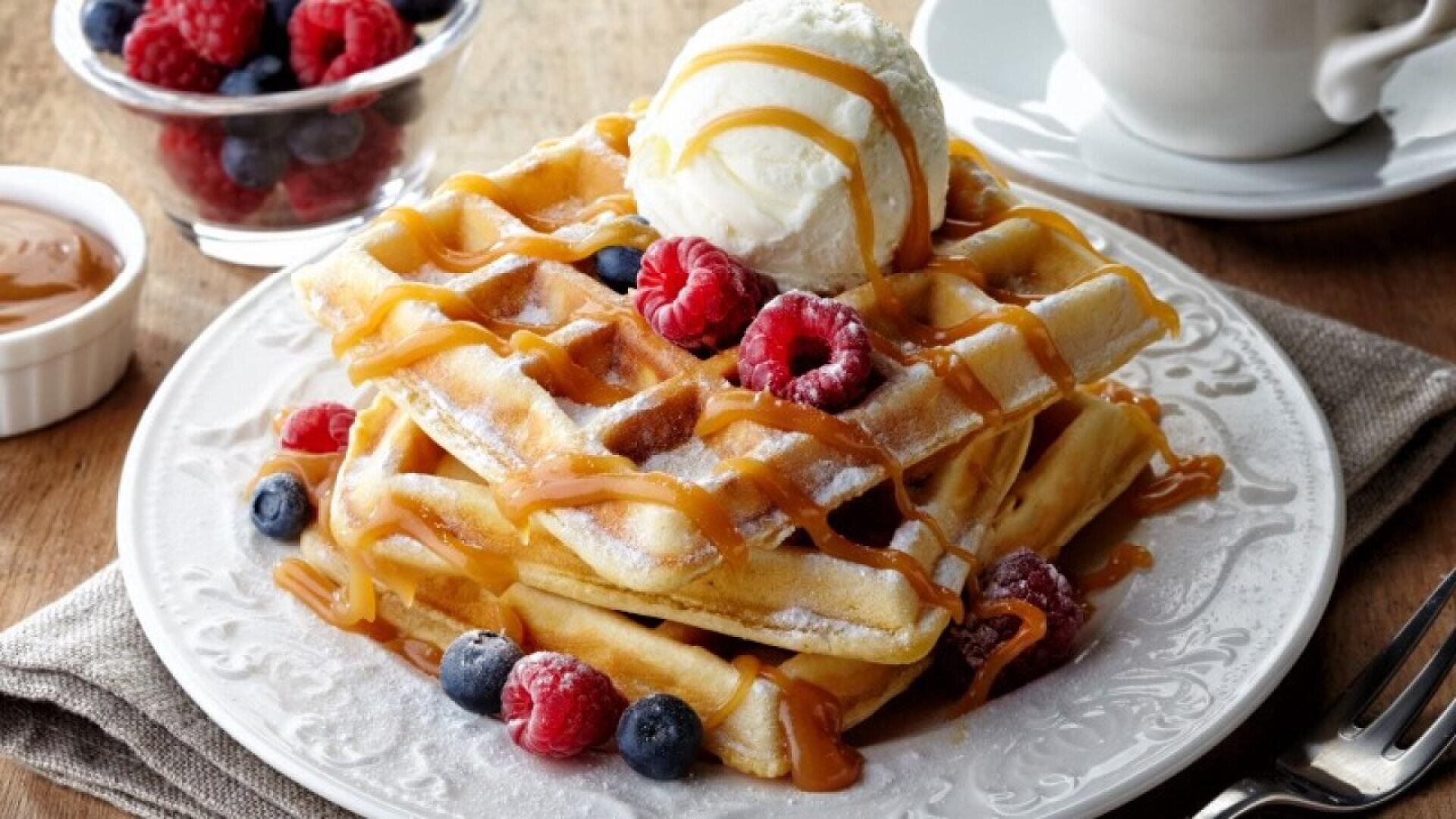 Ein Teller mit Waffeln, belegt mit Vanilleeis, frischen Himbeeren und Heidelbeeren, beträufelt mit Karamellsauce. Im Hintergrund stehen eine kleine Schale mit Beeren und eine Tasse Kaffee.