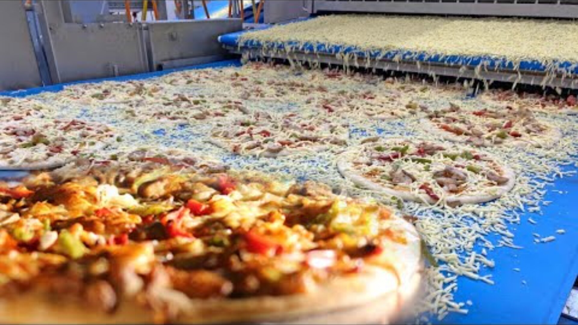 On a blue conveyor belt in a factory, pizzas are topped with grated cheese by a machine, with a close-up of a finished pizza in the foreground.