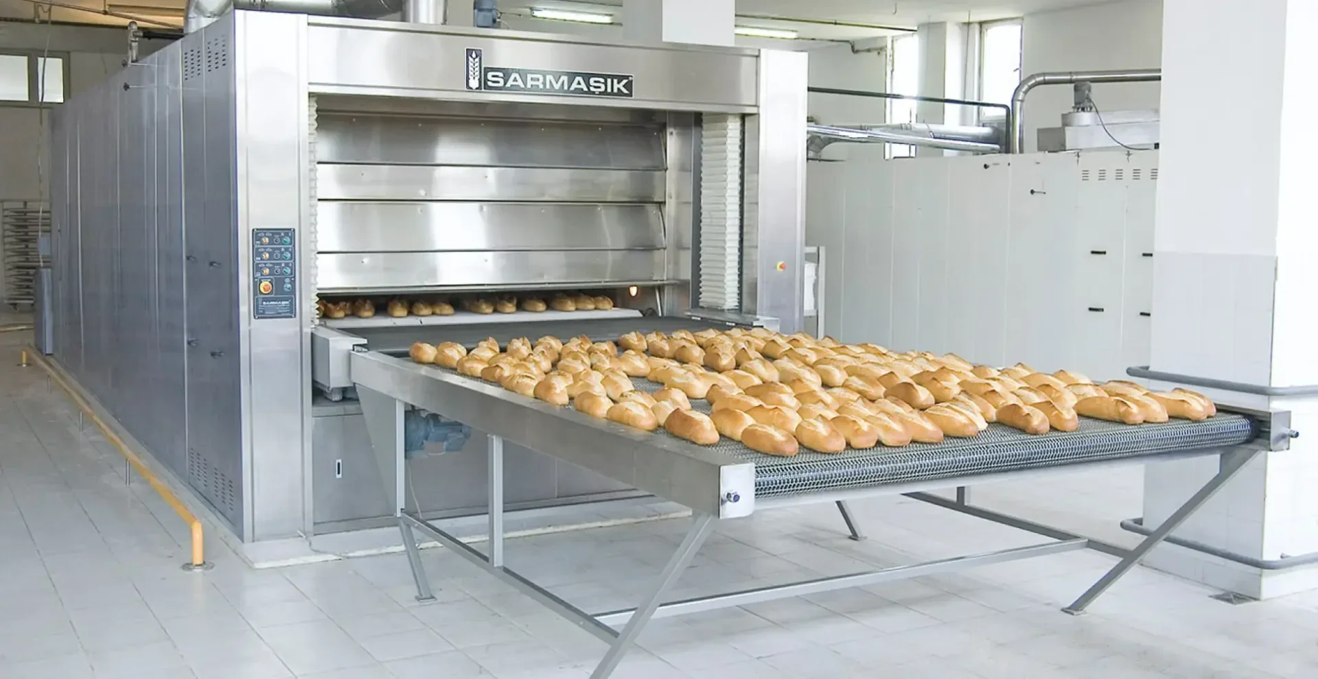 A large industrial bread oven with the brand name “SARMAŞIK” is baking numerous loaves of bread, which are cooling on a conveyor belt in a bright, clean bakery facility.