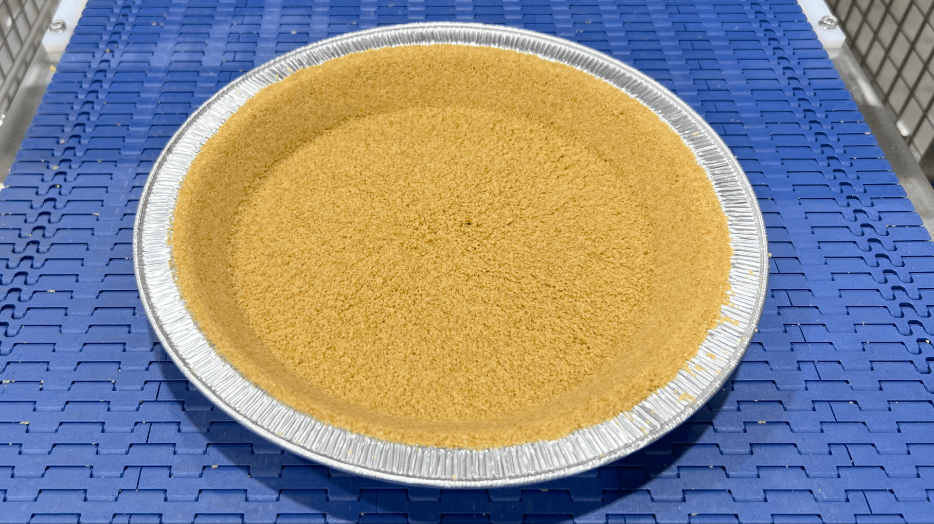 A graham cracker pie crust in a disposable aluminum pan sits on a blue conveyor belt with metal safety railings on each side.