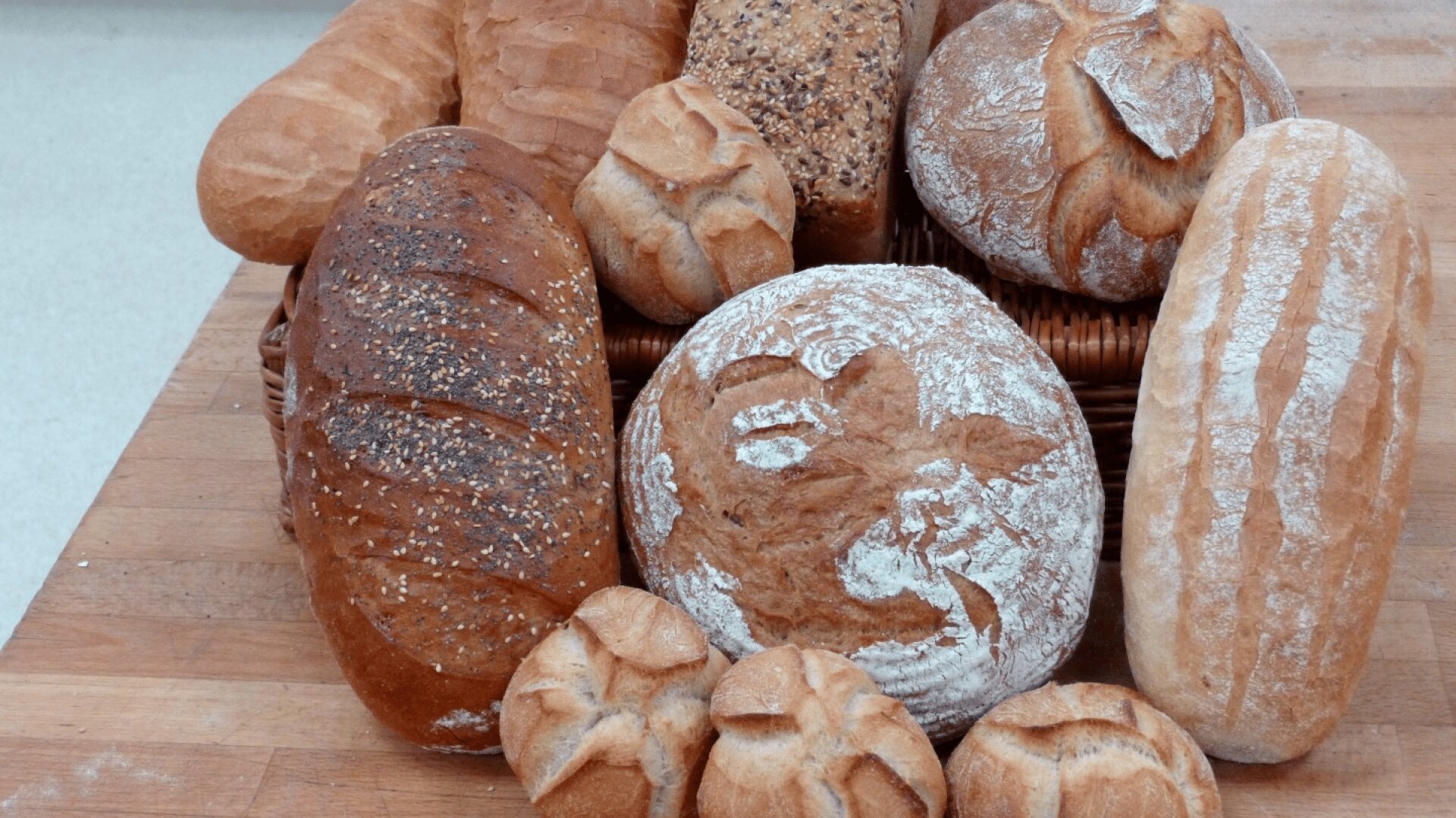 An assortment of artisan breads, including round, oval, and seed-topped loaves, are arranged on a wooden surface and in a wicker basket. The breads vary in color, texture, and crust pattern.