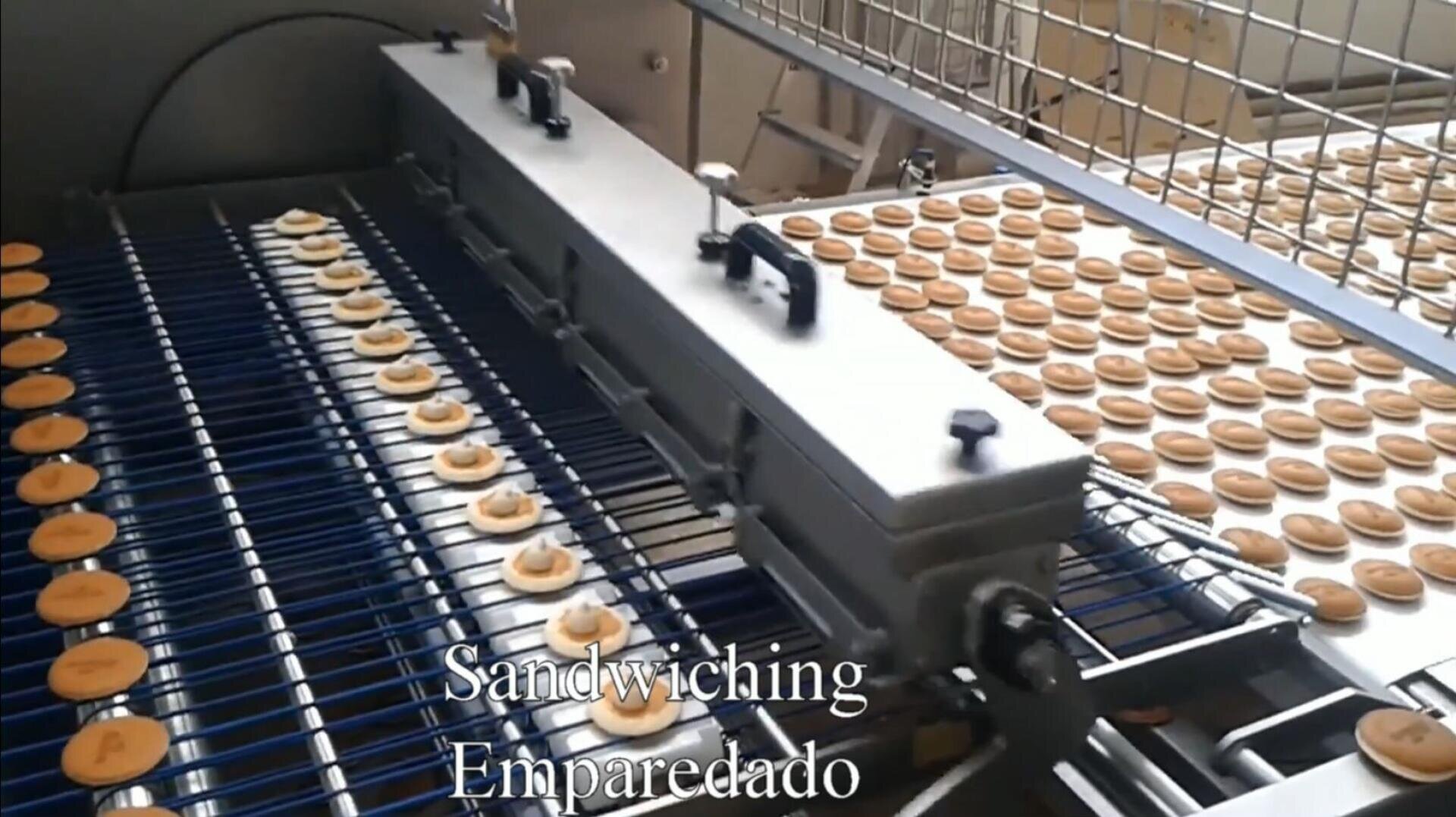 Rows of round cookies move along a conveyor belt under a metal machine in a factory, where cream filling is being added. Text on the image reads “Sandwiching Emparedado.”.