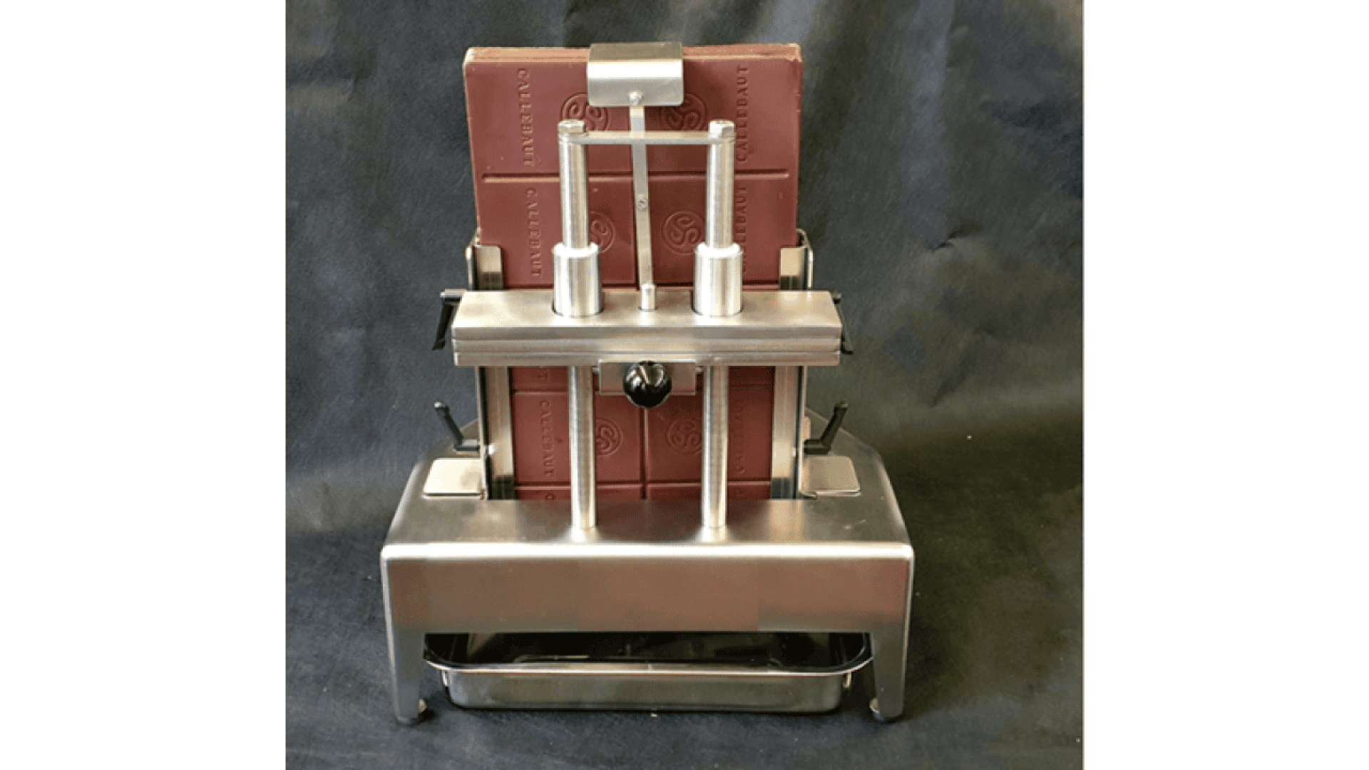 A large chocolate bar held in a stainless steel chocolate cutting machine with three vertical rods and handles, set against a dark background.