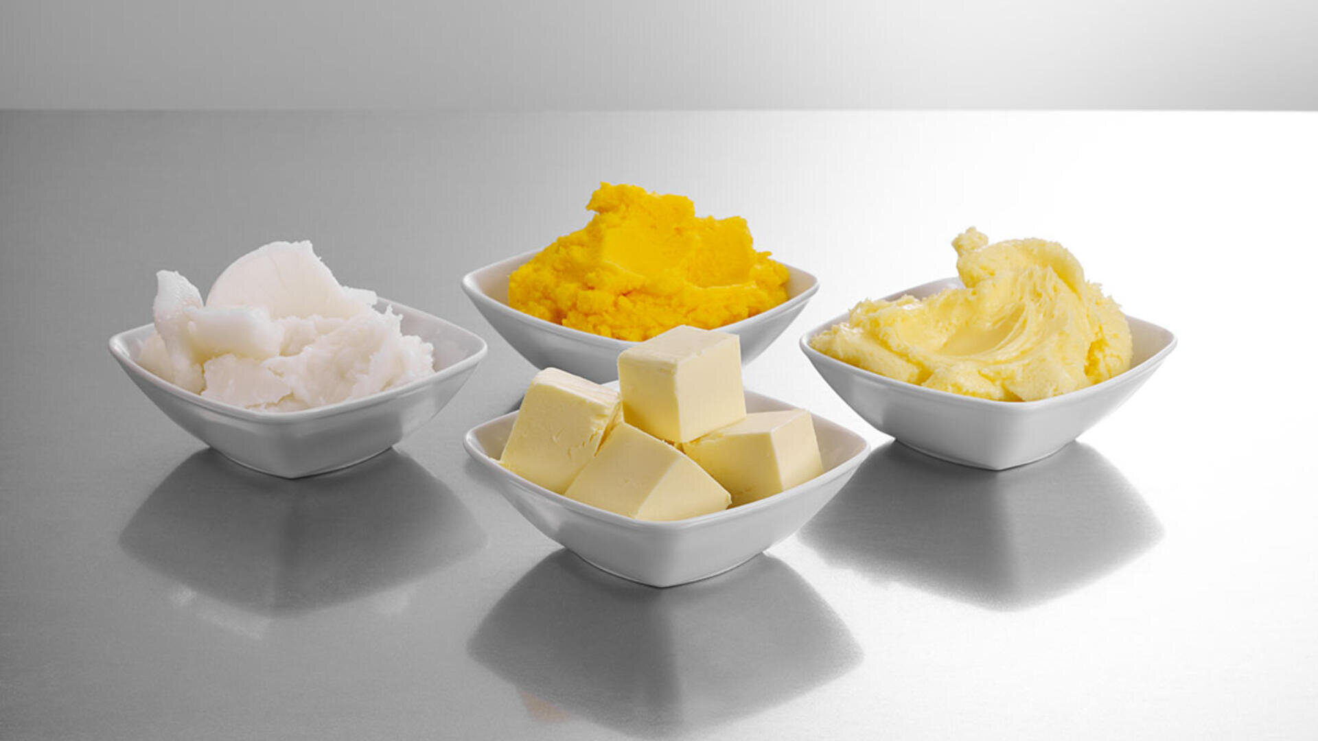 Vier kleine weiße Schälchen, die jeweils eine andere Fettsorte enthalten: weißes Shortening, gelbe Margarine, gewürfelte Butter und cremige hellgelbe Butter, angeordnet auf einer grauen, spiegelnden Oberfläche.