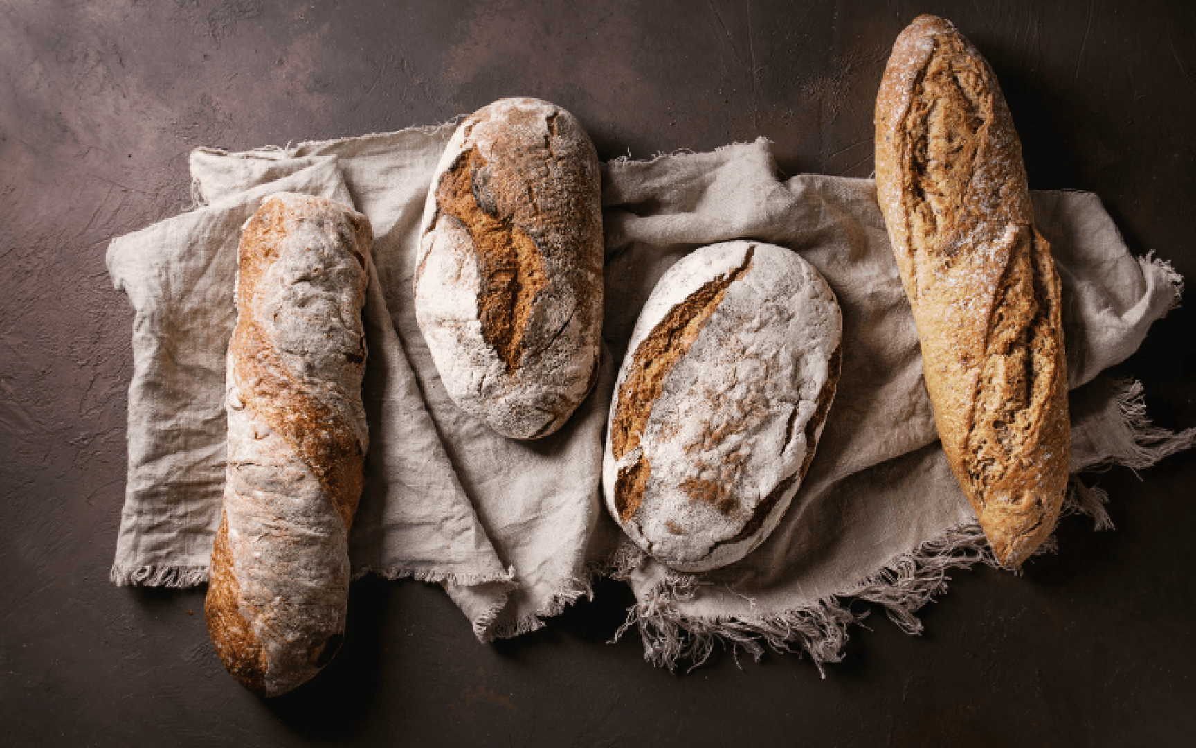 Four rustic loaves of artisanal bread with crusty, golden-brown surfaces rest on a textured, beige cloth atop a dark brown background.