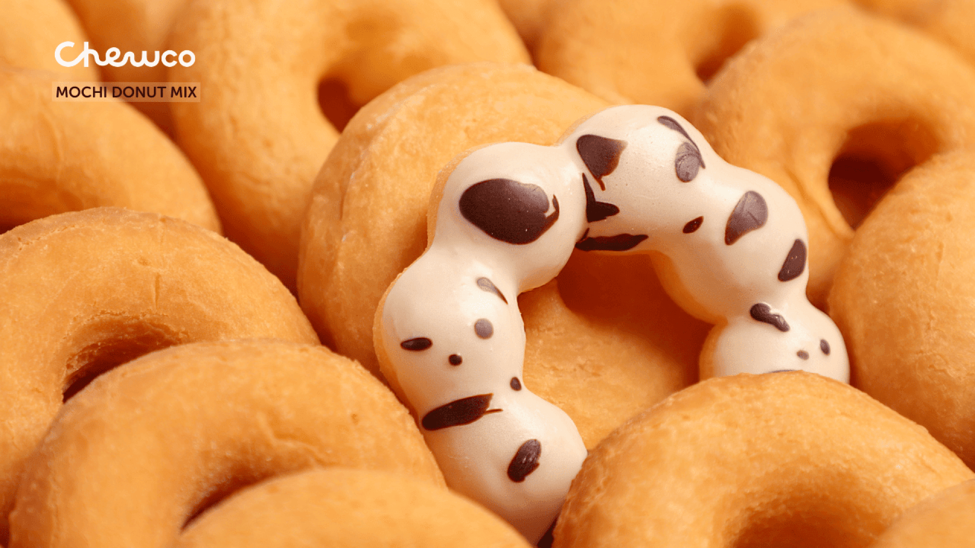Ein weißer Mochi-Donut mit dunklen Flecken liegt auf einem Stapel einfacher goldener Donuts. Das Bild enthält das Logo Chewco und den Text Mochi Donut Mix in der oberen linken Ecke.