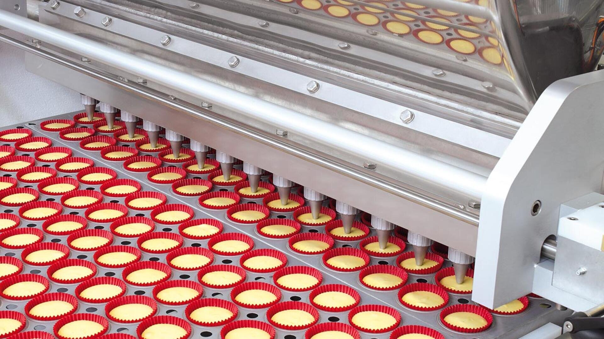 A machine fills rows of red cupcake liners with batter on a conveyor belt in a food production facility, automating the baking preparation process.
