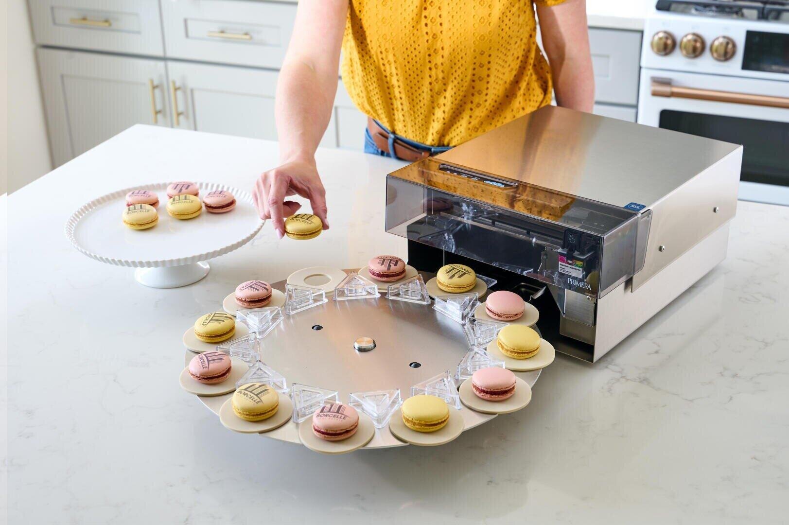Eine Person legt ein Macaron auf ein rotierendes Tablett, das mit einer Maschine verbunden ist, die die Macarons mit Motiven bedruckt. Weitere verzierte und einfarbige Macarons sind auf einem Kuchenständer und dem Tablett in einer modernen Küche zu sehen.