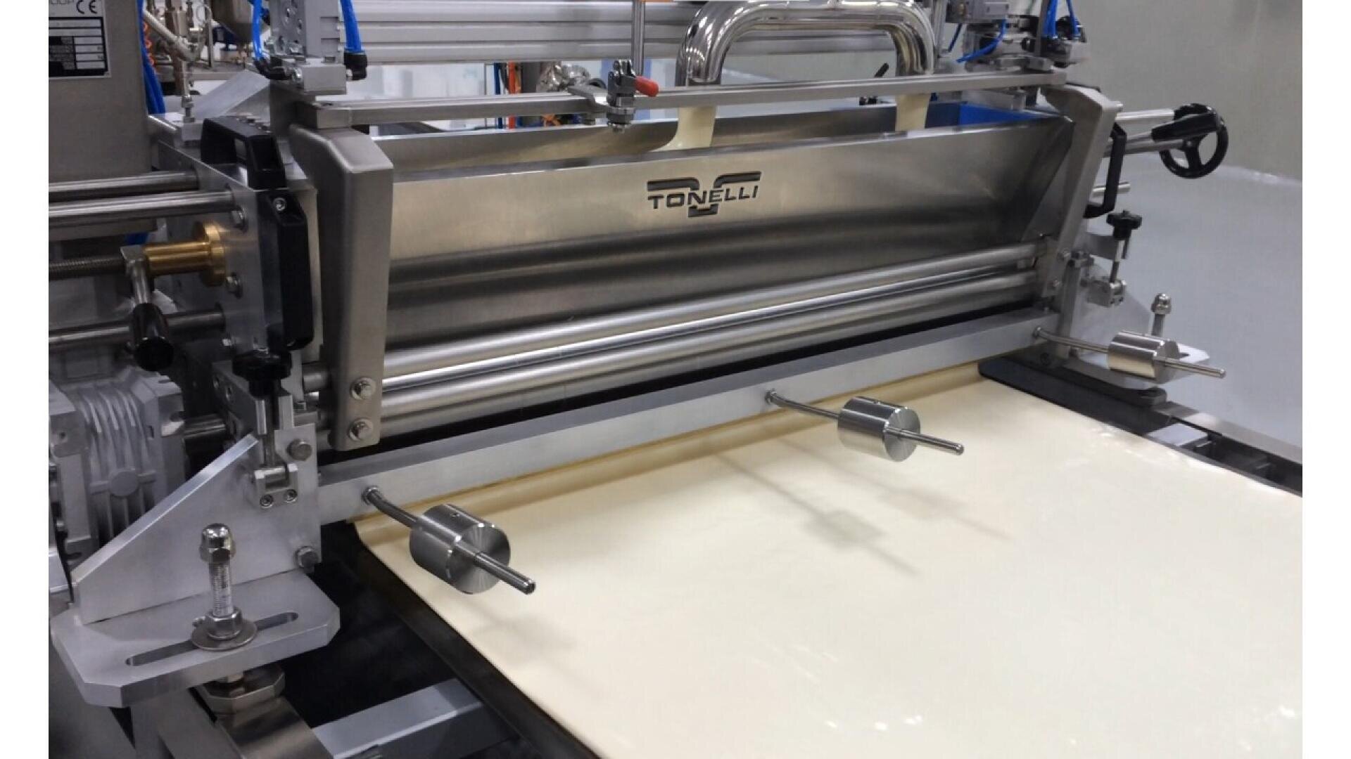 Eine Tonelli-Maschine aus rostfreiem Stahl rollt und glättet Teig auf einem Förderband in einer Lebensmittelverarbeitungsanlage.