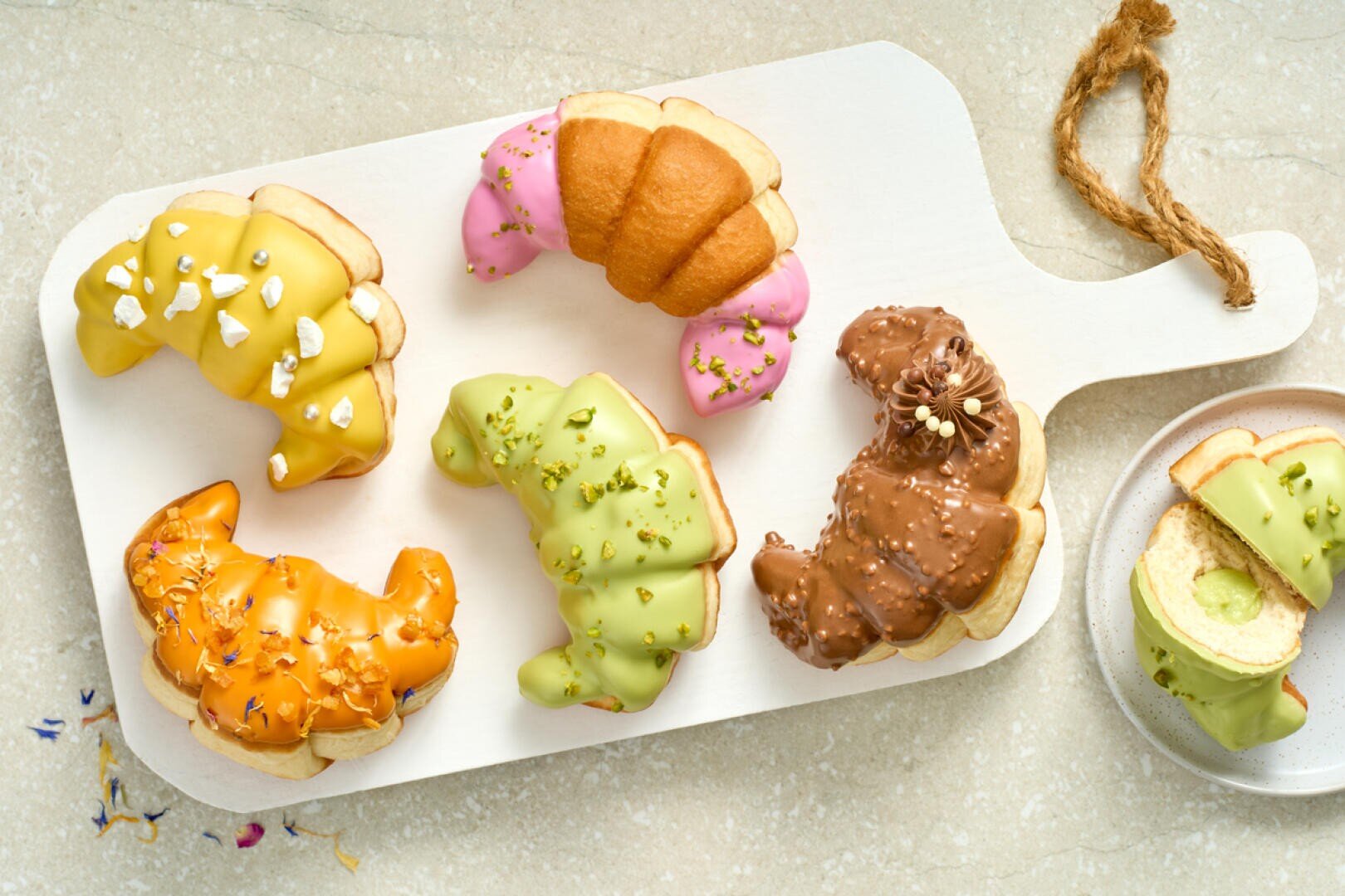 Five colorful croissants with various glazes and toppings are arranged on a white serving board, with one sliced to reveal a creamy filling. The glazes are yellow, pink, green, orange, and chocolate brown.
