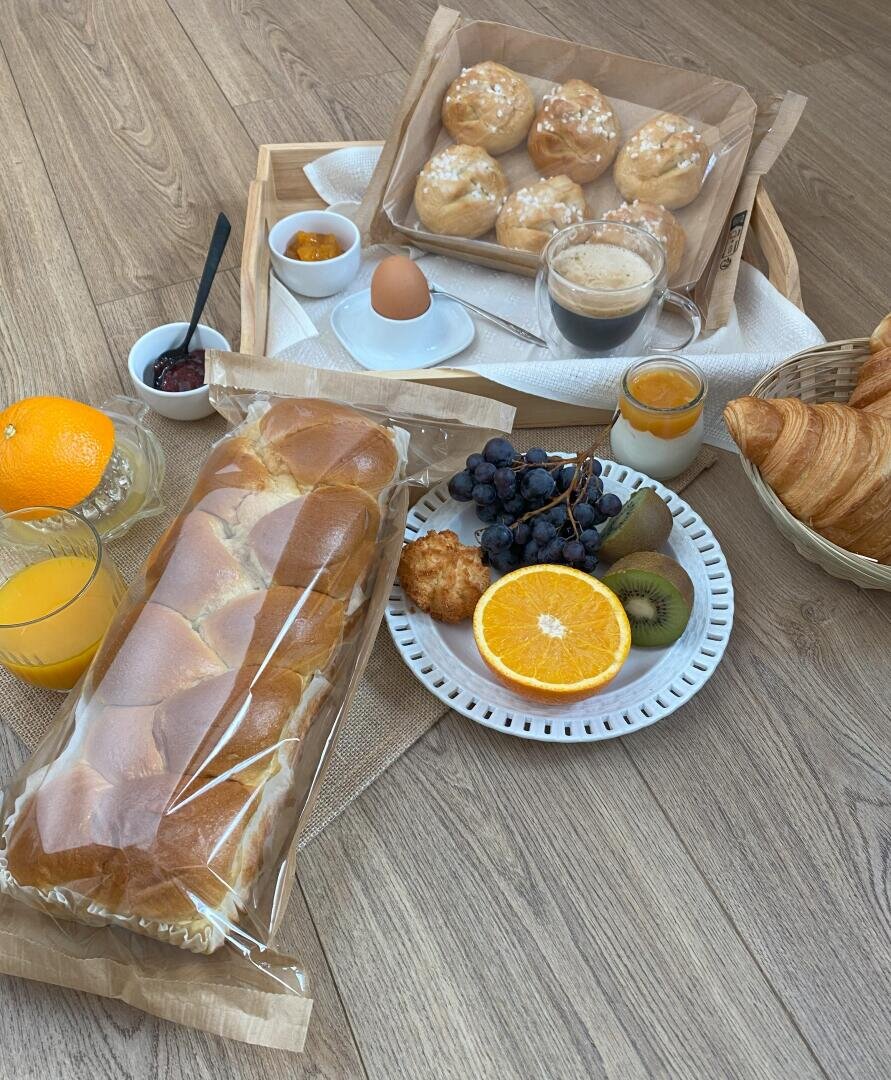 A breakfast spread on a wooden floor with braided bread, orange juice, fruits, baked goods, a soft-boiled egg, jam, and croissants arranged on trays and plates.