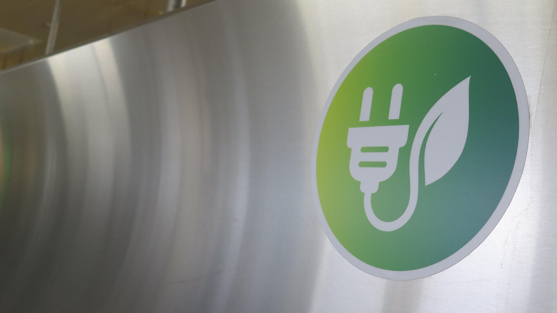 Ein grünes, kreisförmiges Symbol mit einem weißen Stromstecker und einem Blattsymbol, das für umweltfreundliche oder nachhaltige Energie steht, ist auf einer Metalloberfläche abgebildet.